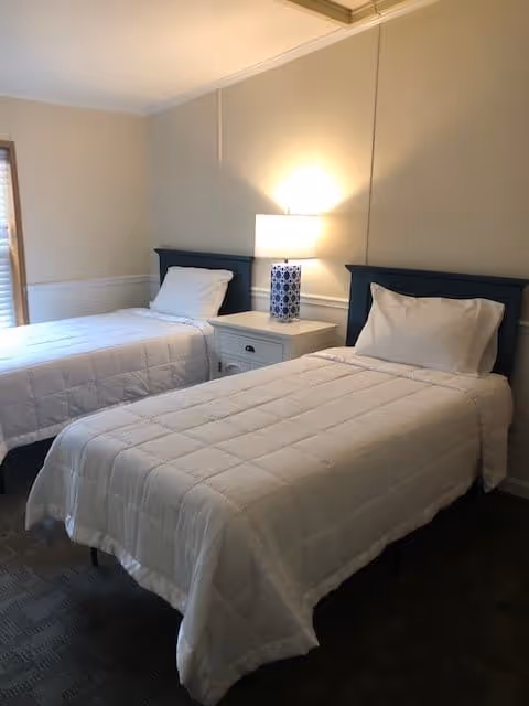 A bedroom with two twin beds, each with white bedding and pillows. Between the beds is a white nightstand with a blue and white patterned lamp. The room has light-colored walls and a window with blinds partially visible on the left side.