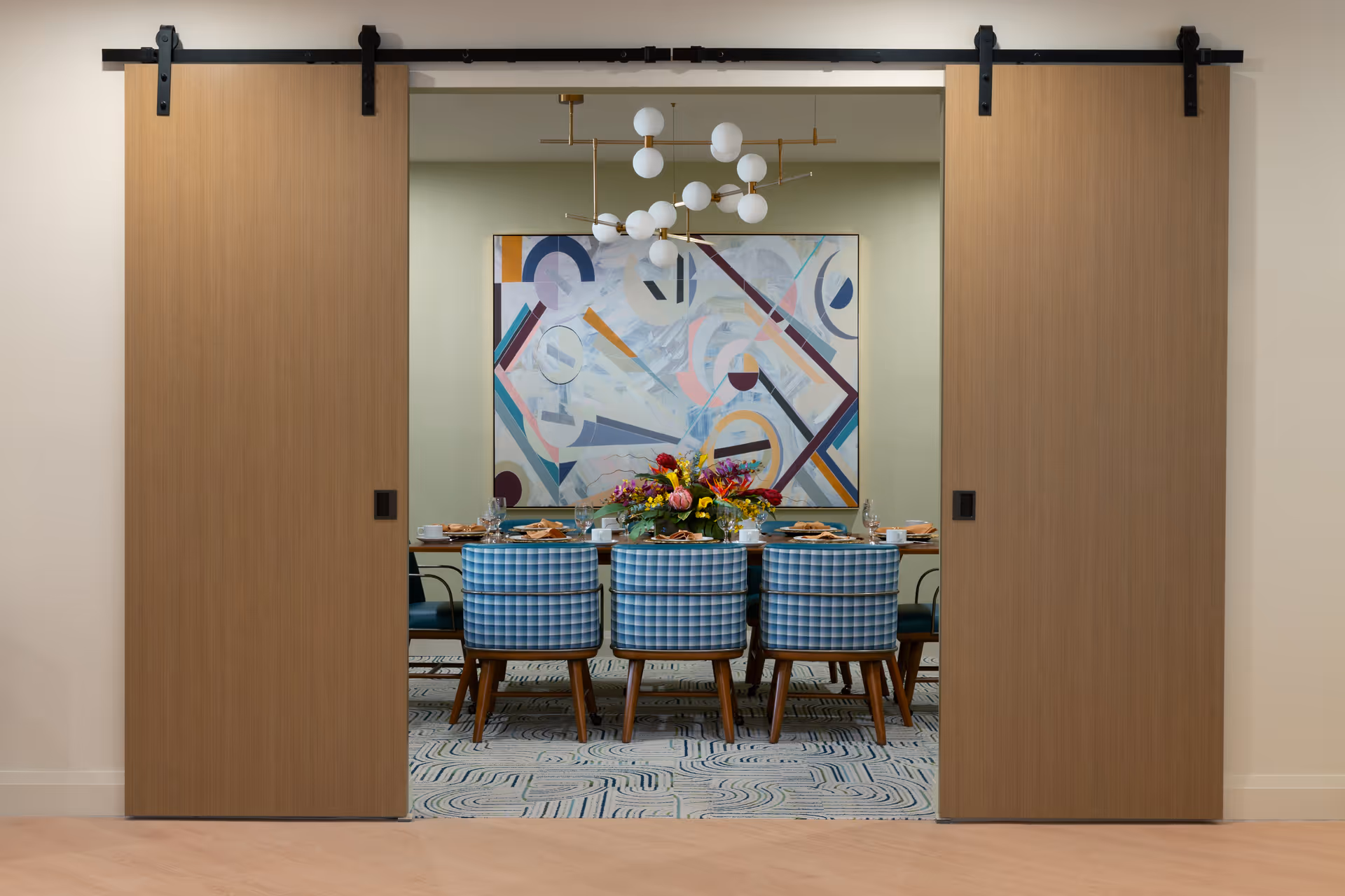 View through partially open wooden sliding doors into a dining room with a table set for a meal. The table is surrounded by six blue checkered chairs, and a colorful floral centerpiece is placed in the middle. A modern chandelier with multiple white globe lights hangs above the table, and an abstract geometric painting is displayed on the far wall.