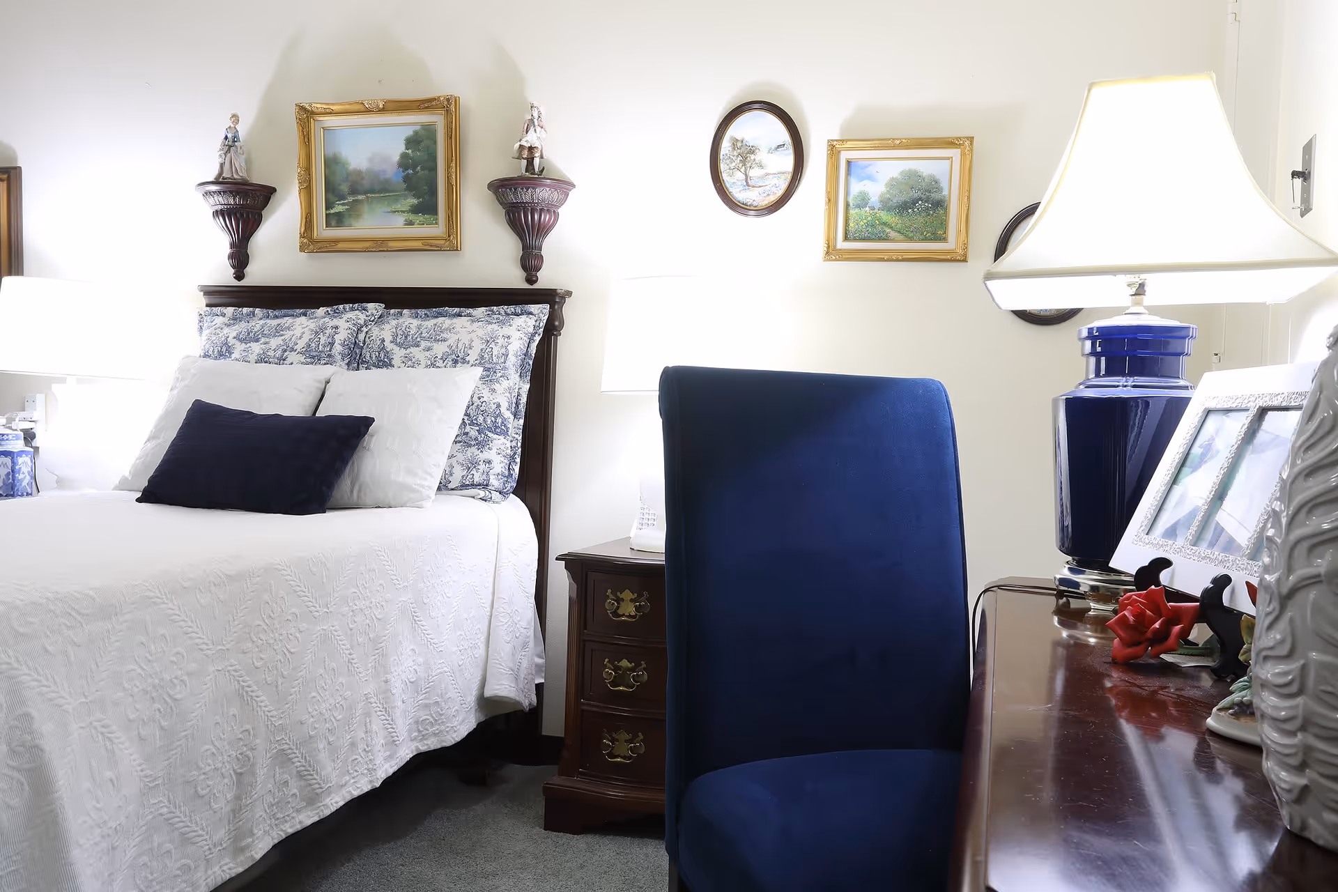 A cozy bedroom featuring a bed with white and blue patterned bedding and multiple pillows. Above the bed are framed landscape paintings and decorative figurines on wall-mounted shelves. To the right of the bed is a wooden nightstand with a lamp. In the foreground, there is a dark blue upholstered chair and a wooden desk with a blue lamp, picture frames, and decorative items.