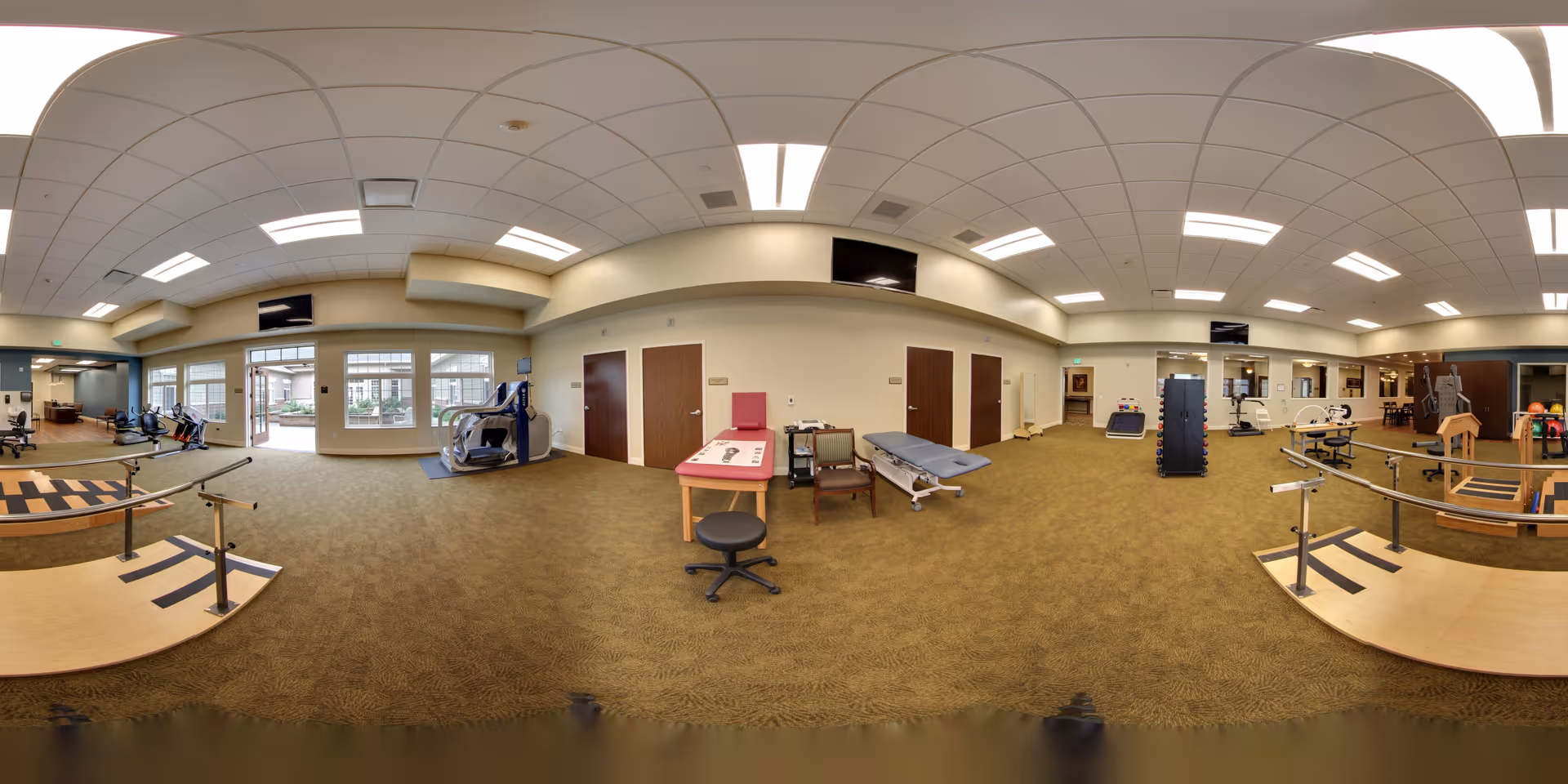Wide panoramic view of a rehabilitation therapy room with exercise equipment, parallel bars for walking practice, therapy tables, chairs, and large windows letting in natural light.