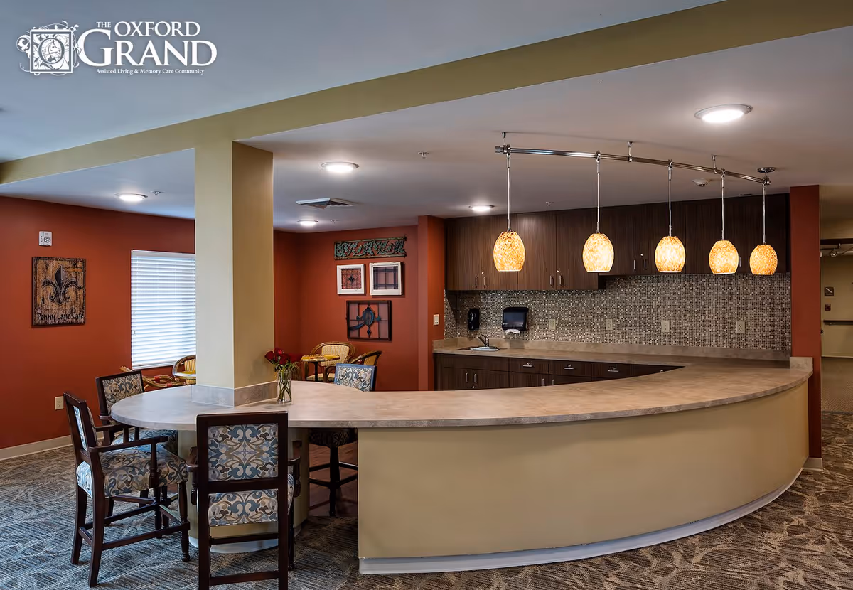 Interior view of a communal area in The Oxford Grand assisted living facility featuring a curved countertop with five hanging pendant lights above it. There are patterned chairs around the counter and a small vase with flowers. The walls are painted in warm tones with decorative artwork, and there is a window with blinds in the background.