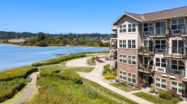 Multi-story senior living building along a grassy waterfront with a paved walkway and seating area under a clear blue sky.