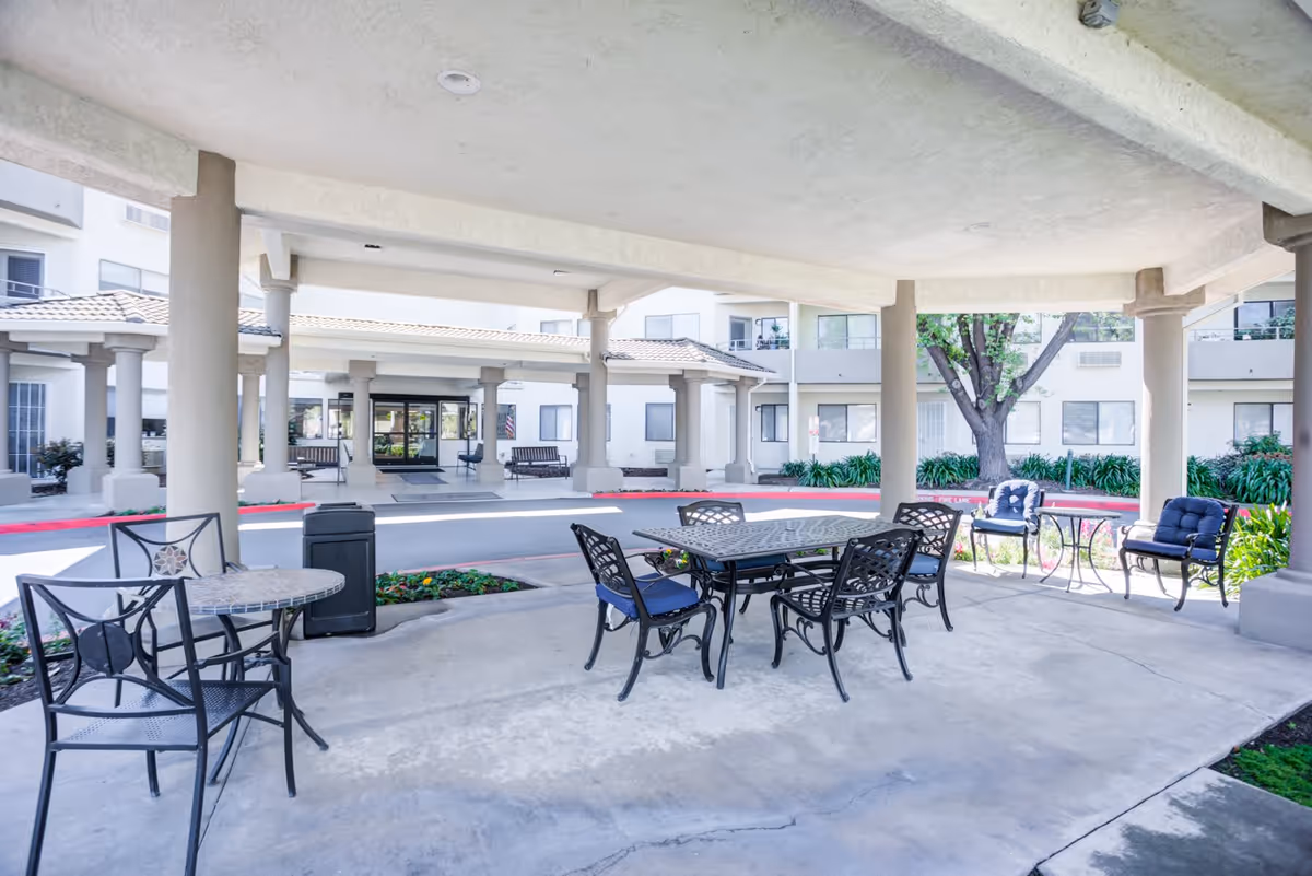 Covered outdoor seating area with metal tables and chairs, some with cushions, located in front of a multi-story building with large windows and a tiled roof. The area is supported by large columns and has a concrete floor, with landscaping and a tree visible in the background.