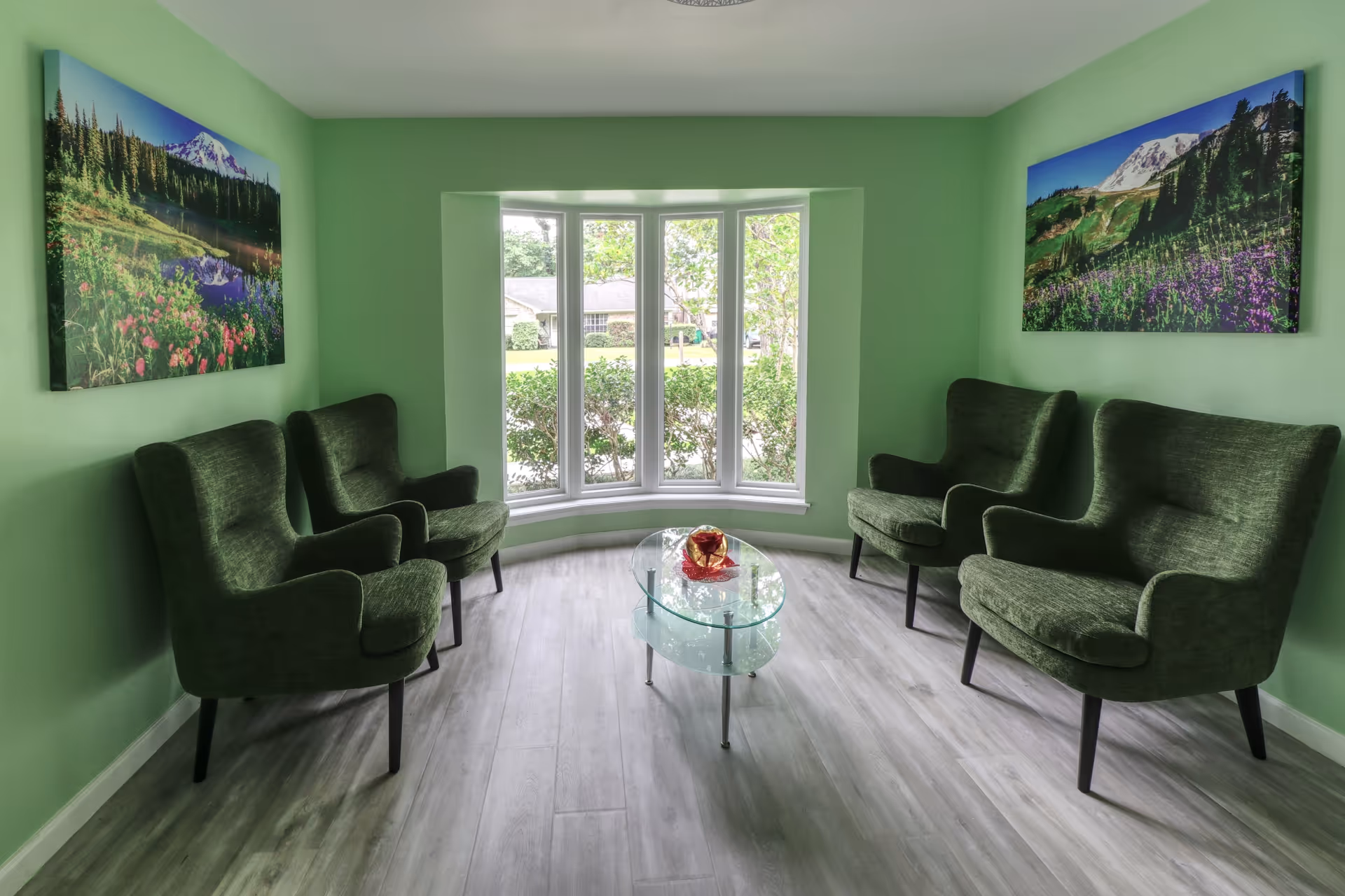 A small sitting area with four green upholstered armchairs arranged around a glass coffee table with a decorative bowl on it. The walls are painted light green and feature two large landscape paintings of mountains and forests. A large bay window lets in natural light and shows greenery outside.