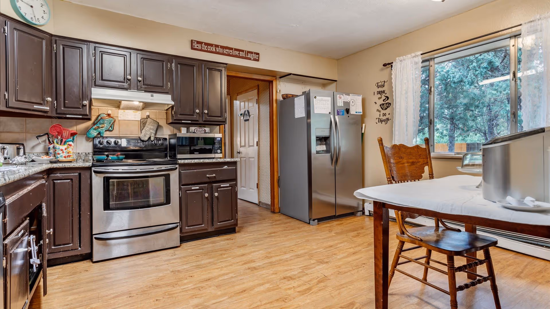 Kitchen with dark cabinets, stainless steel stove and refrigerator, a dining table with chairs, and a large window showing trees outside.