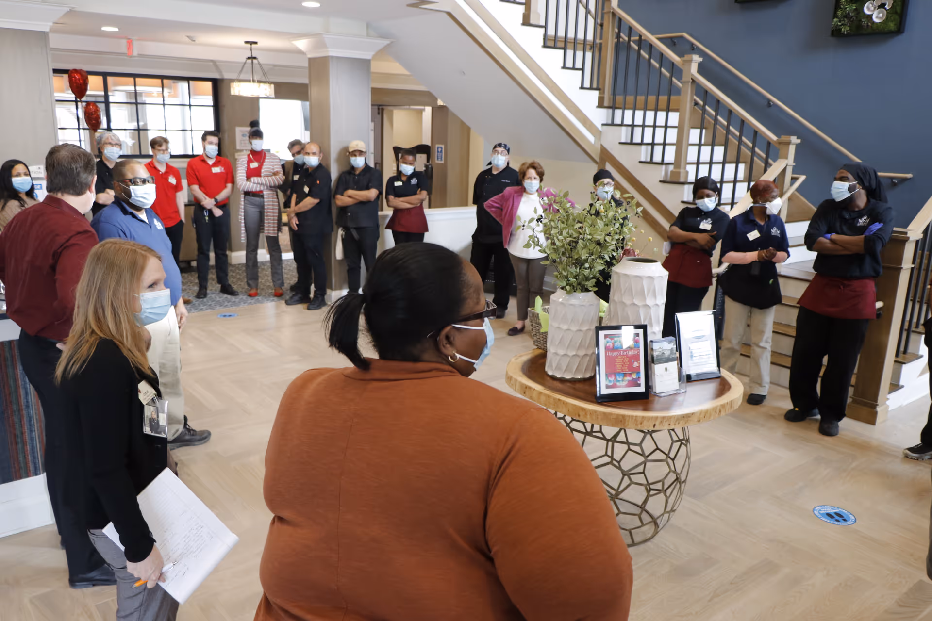 A group of staff members wearing masks standing in a semi-circle inside a senior living facility lobby. The area features a round table with decorative vases and framed pictures, a staircase with wooden railings, and large windows letting in natural light.