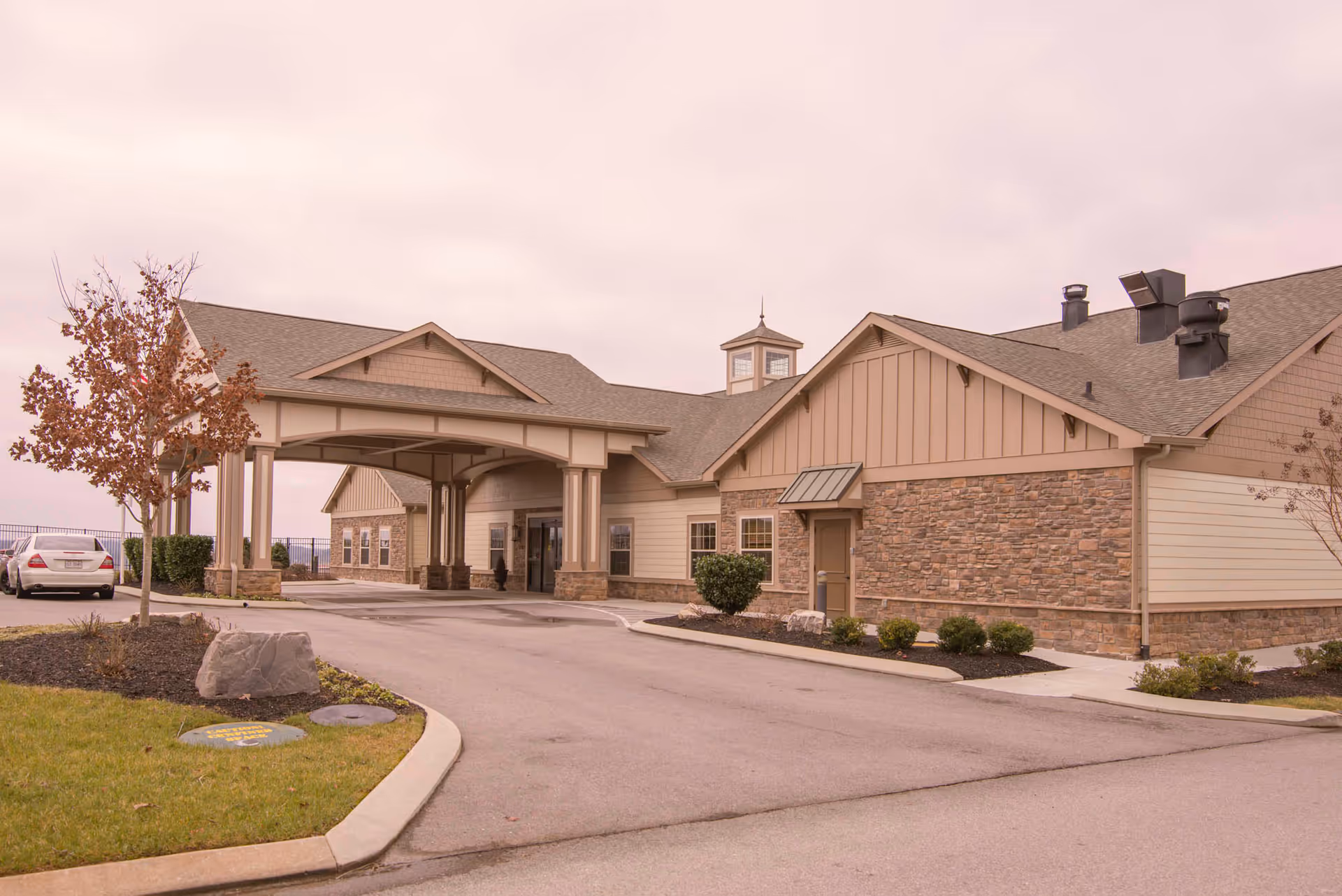 Exterior view of a single-story building with a covered entrance driveway, beige siding, stone accents, and a small cupola on the roof. There is a car parked on the left side near a landscaped area with a tree and bushes.