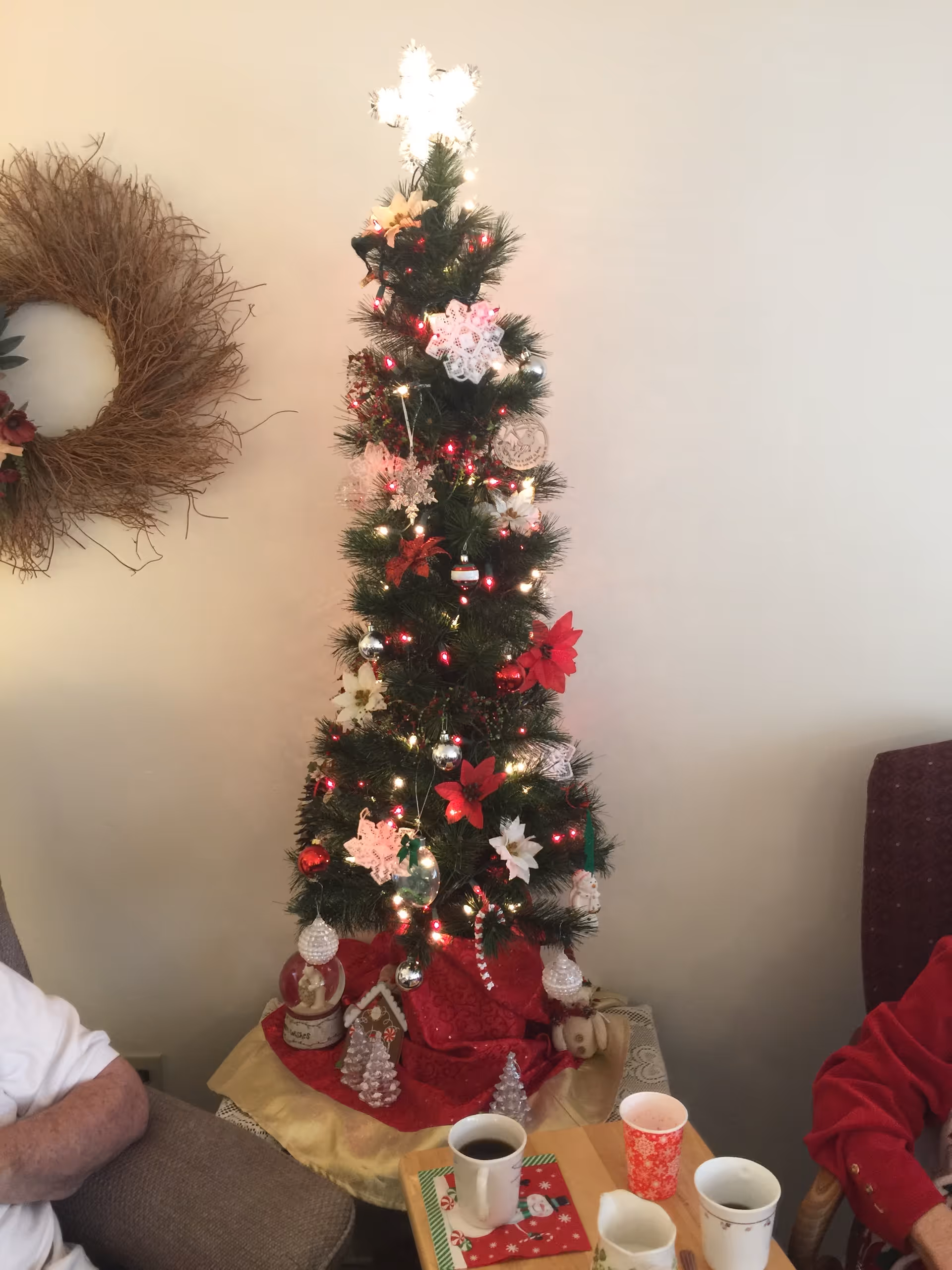 A small decorated Christmas tree with lights and ornaments next to chairs and a table holding cups.