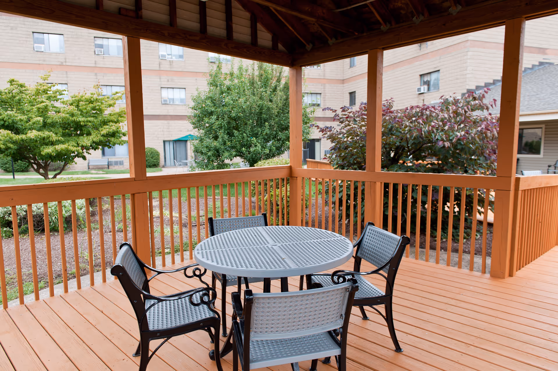 Covered outdoor patio area with a round metal table and four matching chairs. The patio has wooden railings and flooring, surrounded by green bushes and trees. In the background, there is a multi-story building with windows and air conditioning units.