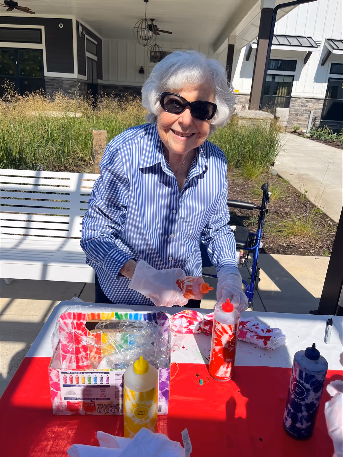 An elderly woman with white hair and sunglasses is smiling while standing at a table outdoors. She is wearing a blue and white striped shirt and white gloves, holding a bottle of orange dye. On the table are bottles of yellow, red, and blue dye, a tie-dye kit box, and white fabric items being tie-dyed. The setting appears to be a patio area with a white bench, greenery, and a building in the background.
