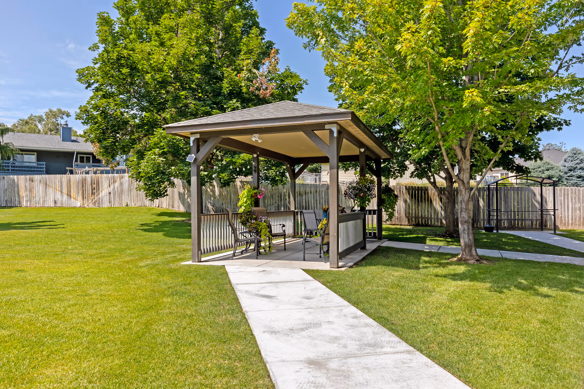 A small outdoor gazebo with a roof and open sides, containing benches and chairs, situated on a concrete path surrounded by green grass and trees in a fenced yard under a clear blue sky.