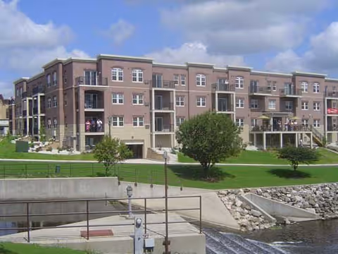 A large, multi-story brick residential building with balconies overlooking a grassy area and a small waterway with a concrete embankment. The sky is partly cloudy.