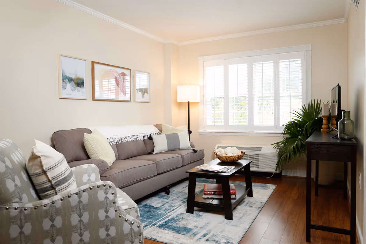 Sunlit living room with a gray sofa, patterned armchair, coffee table, floor lamp, and a window with shutters.