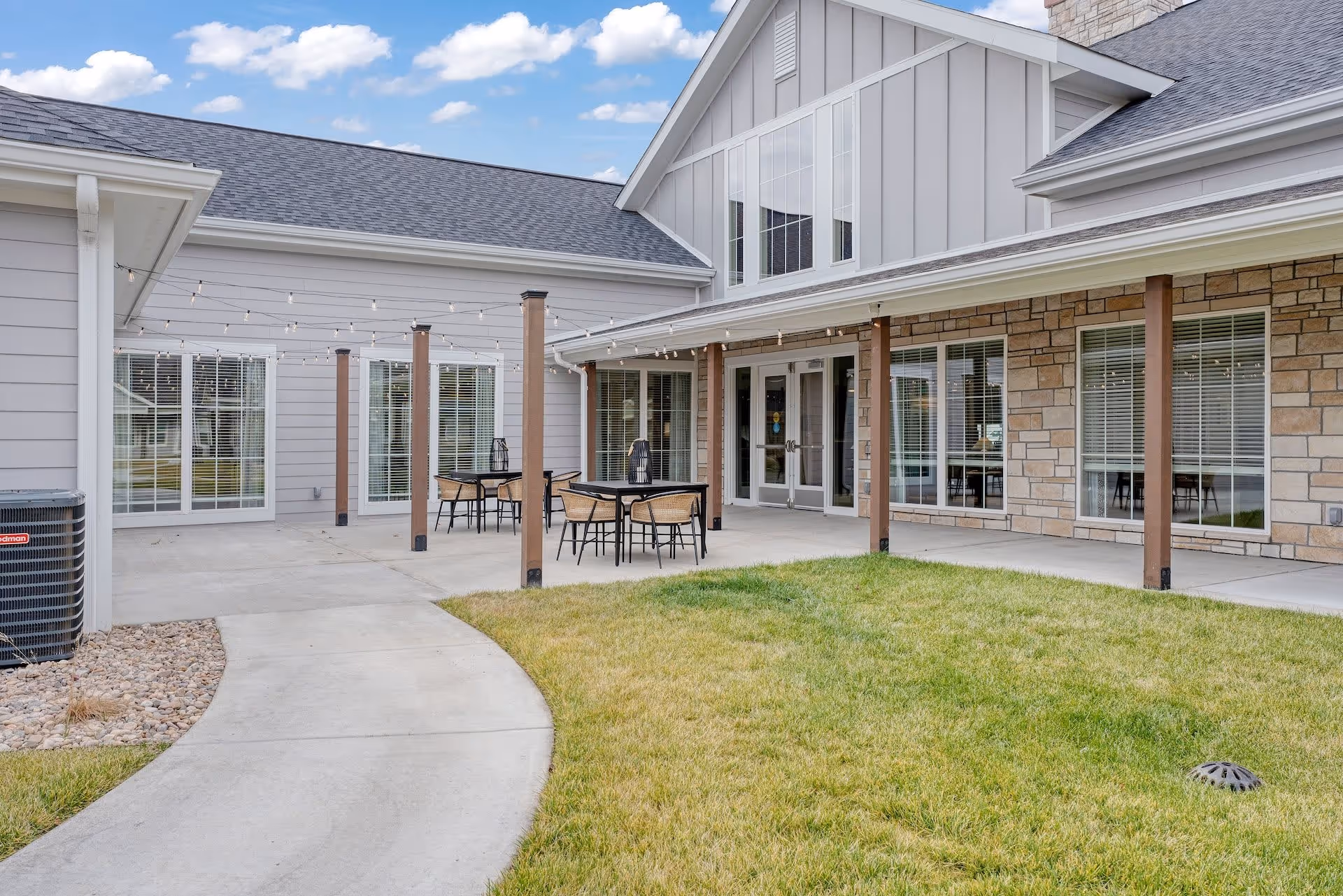 Exterior courtyard and covered patio of a senior living building with tables, string lights, and large windows.