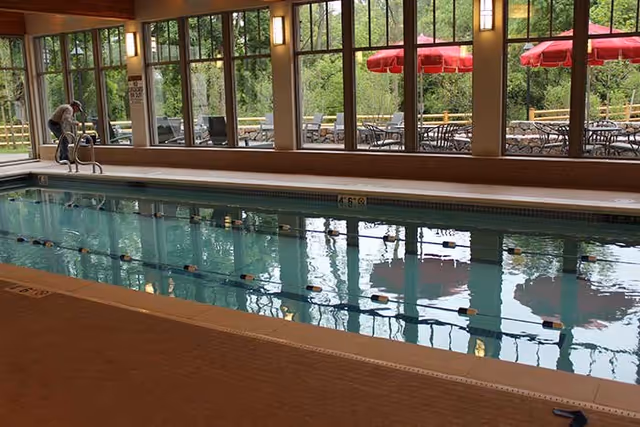 Indoor lap pool with lane dividers and large windows overlooking an outdoor patio with red umbrellas.