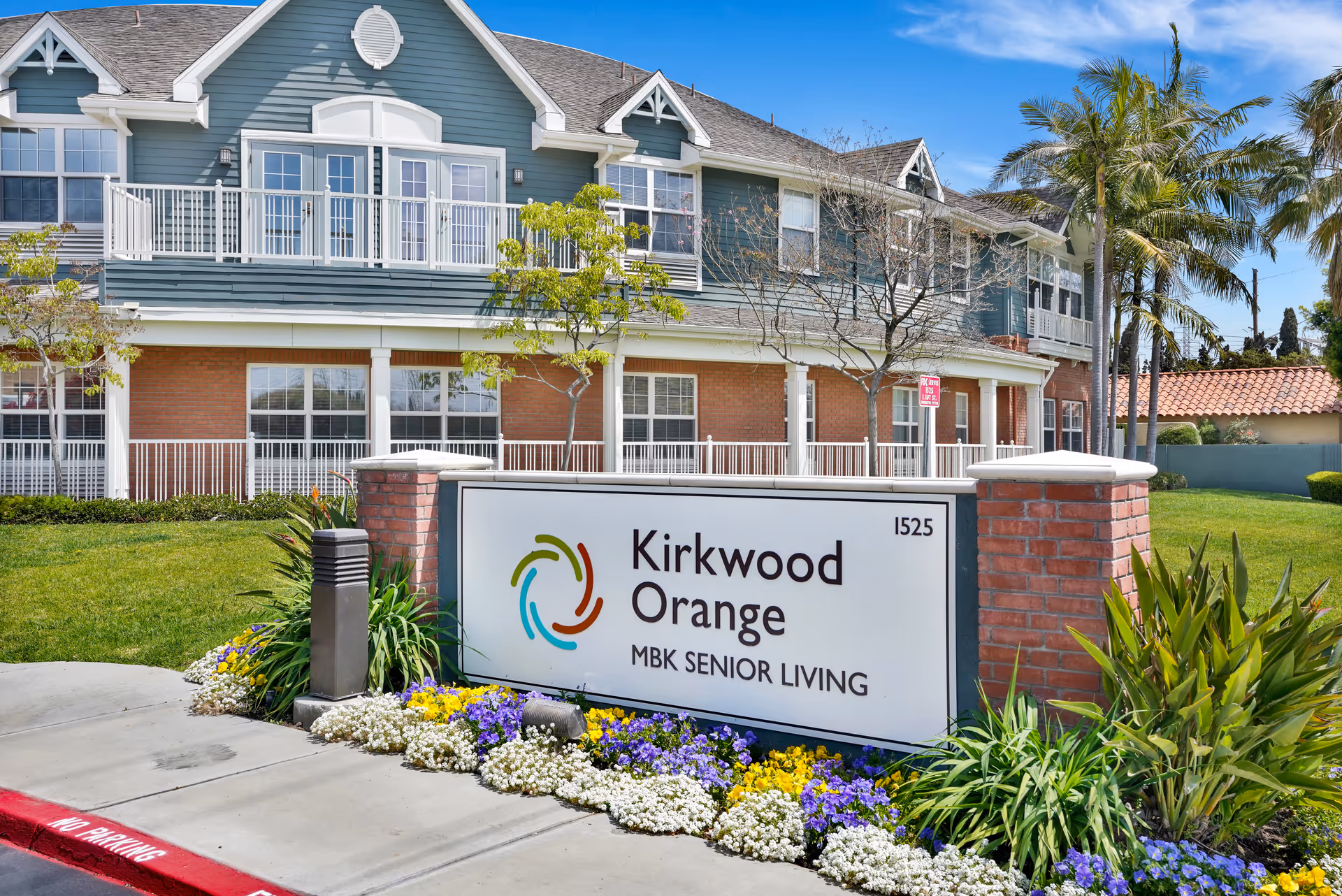Exterior view of Kirkwood Orange senior living facility with a large sign in front displaying the facility name and surrounded by colorful flowers and greenery. The building has a blue and brick facade with white railings and multiple windows under a clear blue sky.