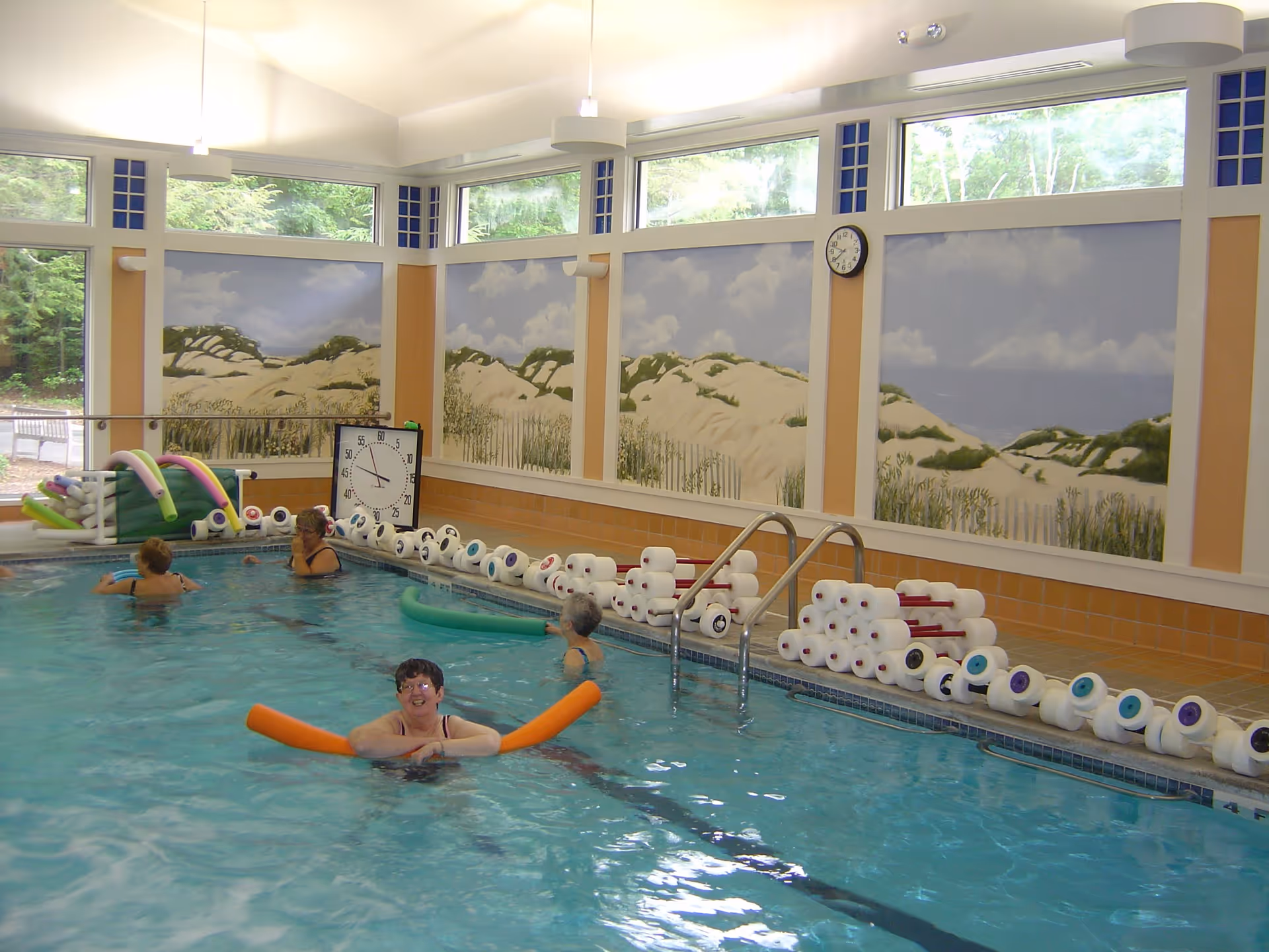 Indoor swimming pool with several elderly people enjoying water exercise. The pool area has large windows near the ceiling letting in natural light and walls decorated with murals of sand dunes and grass. Pool noodles and foam weights are stacked along the poolside, and a large clock is mounted on the wall.