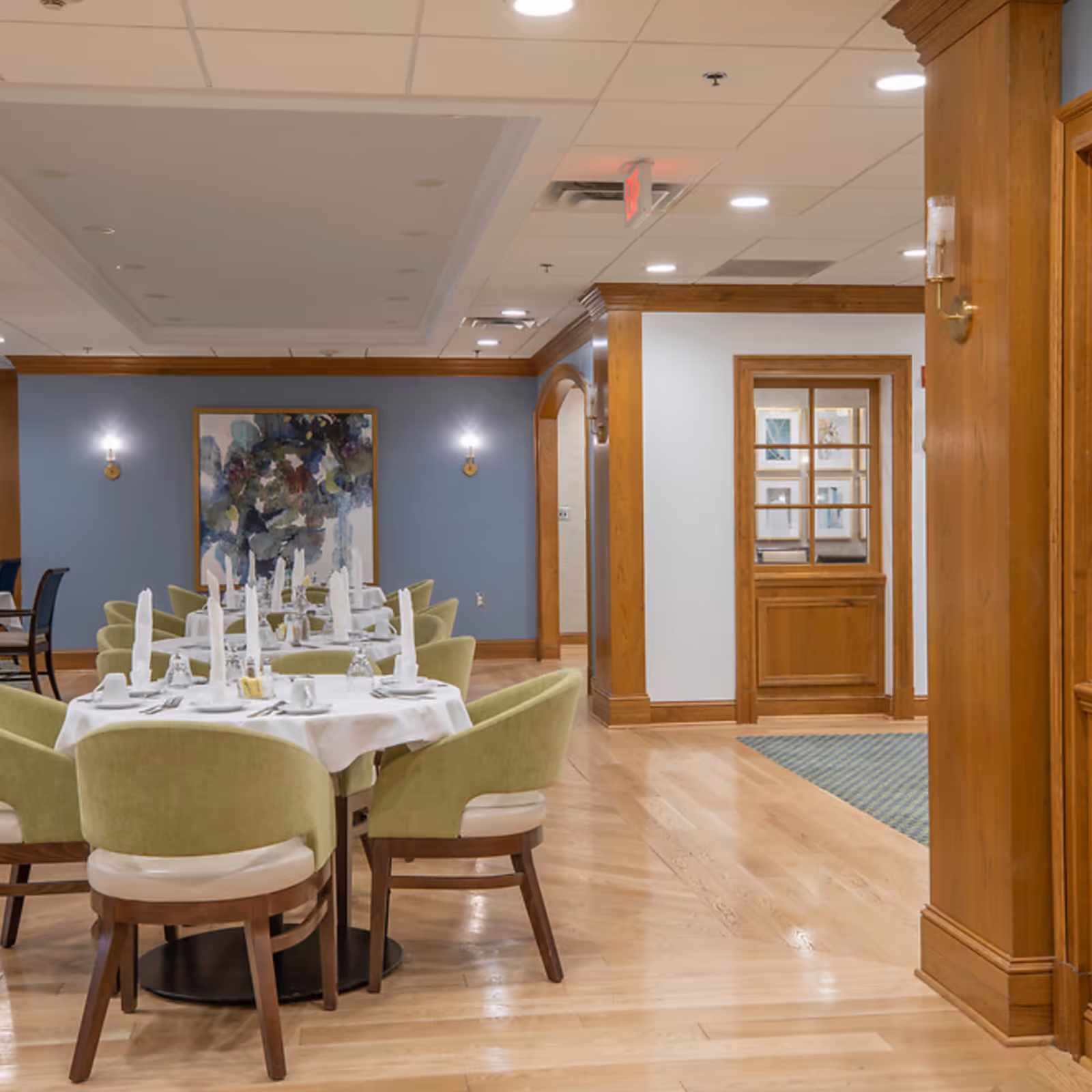 A dining room with round tables covered in white tablecloths, set with white napkins, cups, and silverware. The chairs around the tables have light green upholstery and wooden legs. The room features wooden trim, light hardwood floors, blue and white walls, and a large abstract painting on the far wall. Recessed ceiling lights illuminate the space.