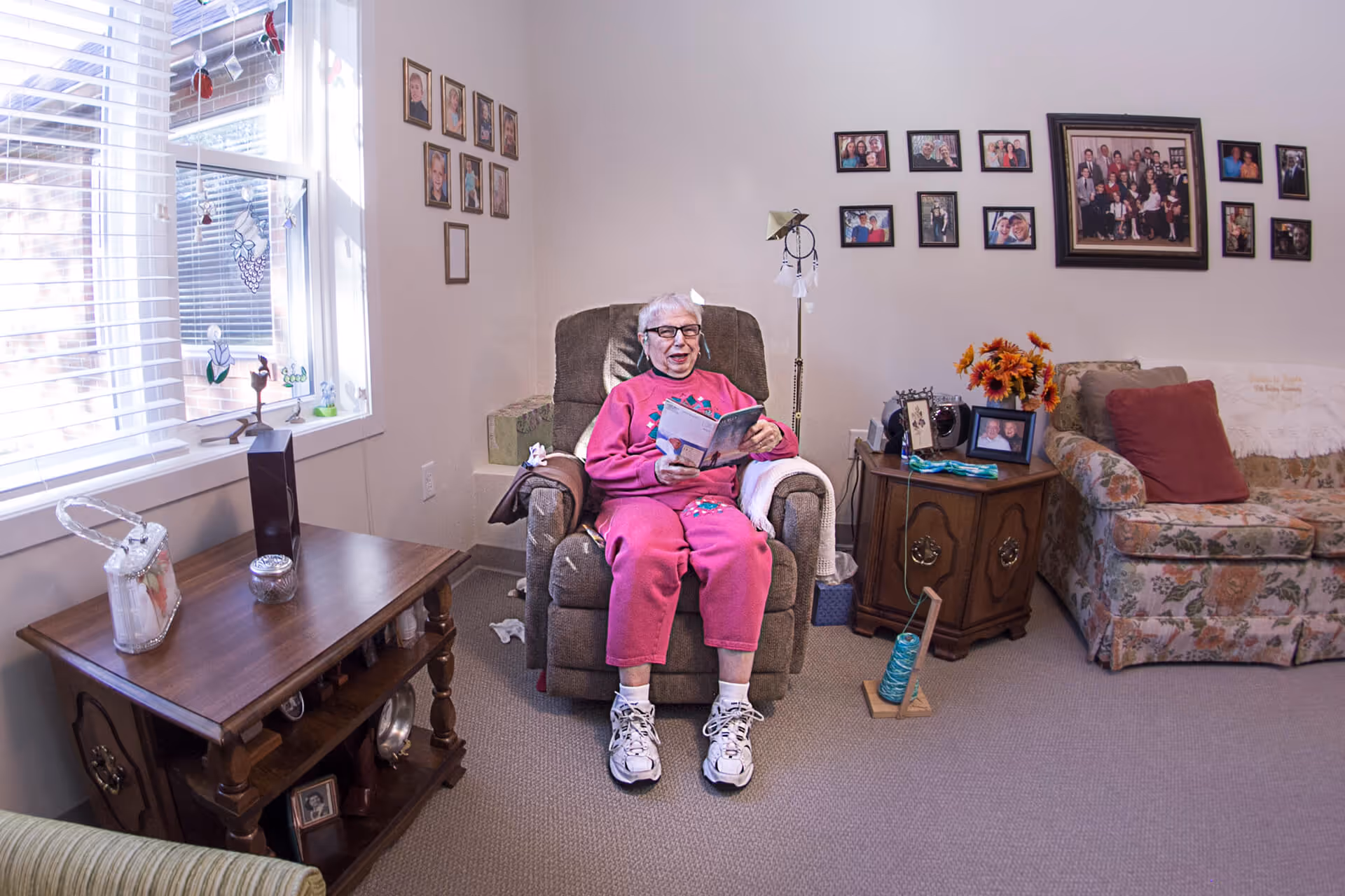 An elderly woman wearing a pink outfit and white sneakers is sitting in a brown recliner chair in a cozy living room. She is holding and reading a book. The room has a window with blinds on the left, a wooden side table with decorative items, and a floral-patterned couch with a red pillow on the right. The walls are decorated with multiple framed family photos.