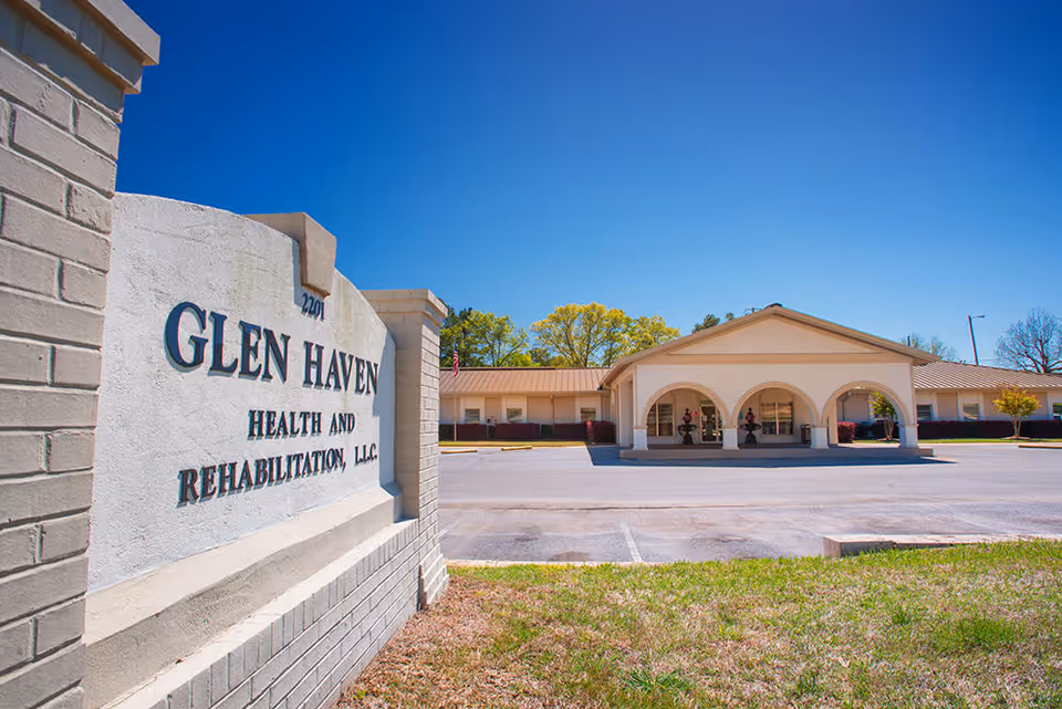 Exterior view of Glen Haven Health and Rehabilitation, LLC building with a large sign in the foreground displaying the facility's name. The building has a covered entrance with three arches and a parking area in front, under a clear blue sky.
