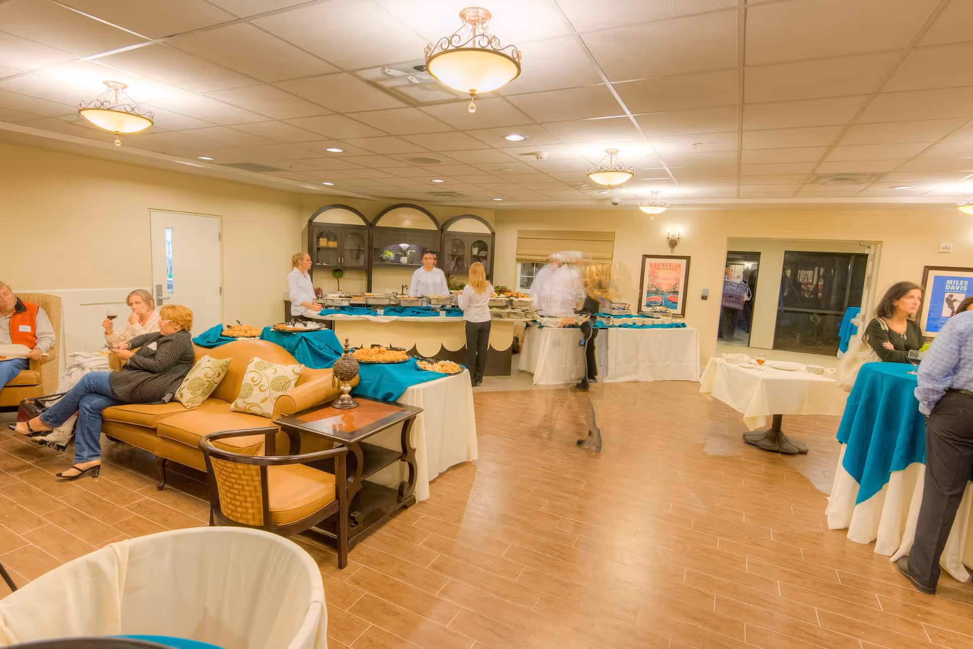 Residents and staff mingling around buffet tables in a well-lit community room with seating and teal tablecloths.
