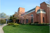 Brick senior living building with manicured lawn, blooming tree, and clear blue sky.