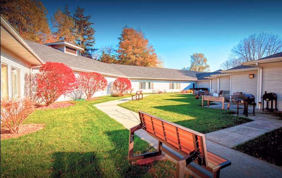 Outdoor courtyard area at Corridor Crossing Place with green grass, a concrete walkway, red-leaved bushes, several benches, and multiple barbecue grills. The courtyard is surrounded by single-story building walls under a clear blue sky.