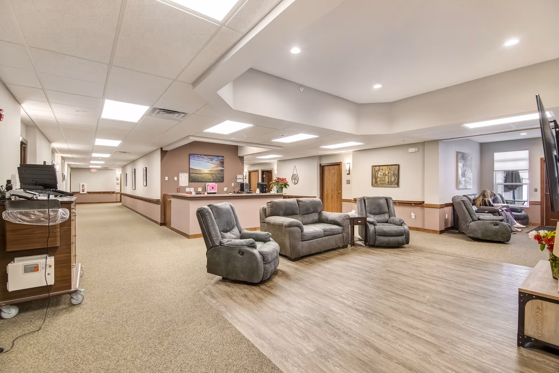 A spacious senior living facility common area with gray recliner chairs and a loveseat arranged on a wood floor section. The area is well-lit with ceiling lights and has beige walls with wooden trim. A hallway extends to the left with framed pictures on the walls. In the background, two people are seated on recliners near a window. A reception desk with a flower pot and computer is visible behind the seating area.