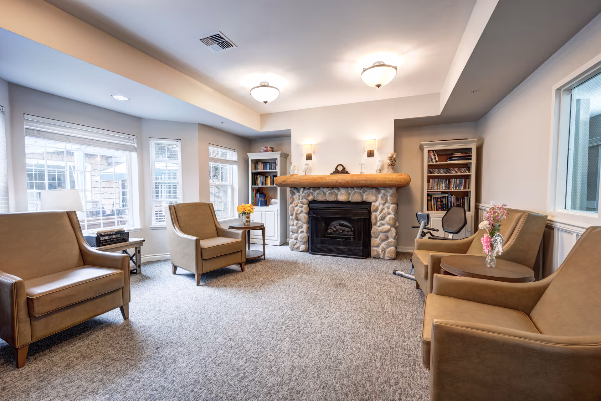 A cozy living room with four tan armchairs arranged around a stone fireplace with a wooden mantle. Two white bookshelves filled with books flank the fireplace. There are windows on the left side letting in natural light, and small tables with flower vases next to the chairs. The room has beige carpet and soft overhead lighting.