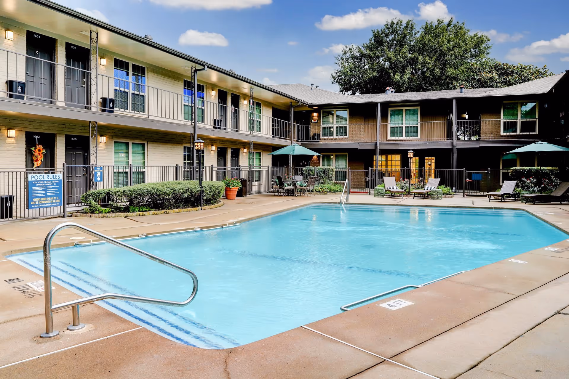 Outdoor swimming pool area in a senior living facility with a two-story building surrounding the pool. The building has multiple doors and windows, some with potted plants and patio furniture including tables with umbrellas and lounge chairs around the pool. The sky is clear with a few clouds.