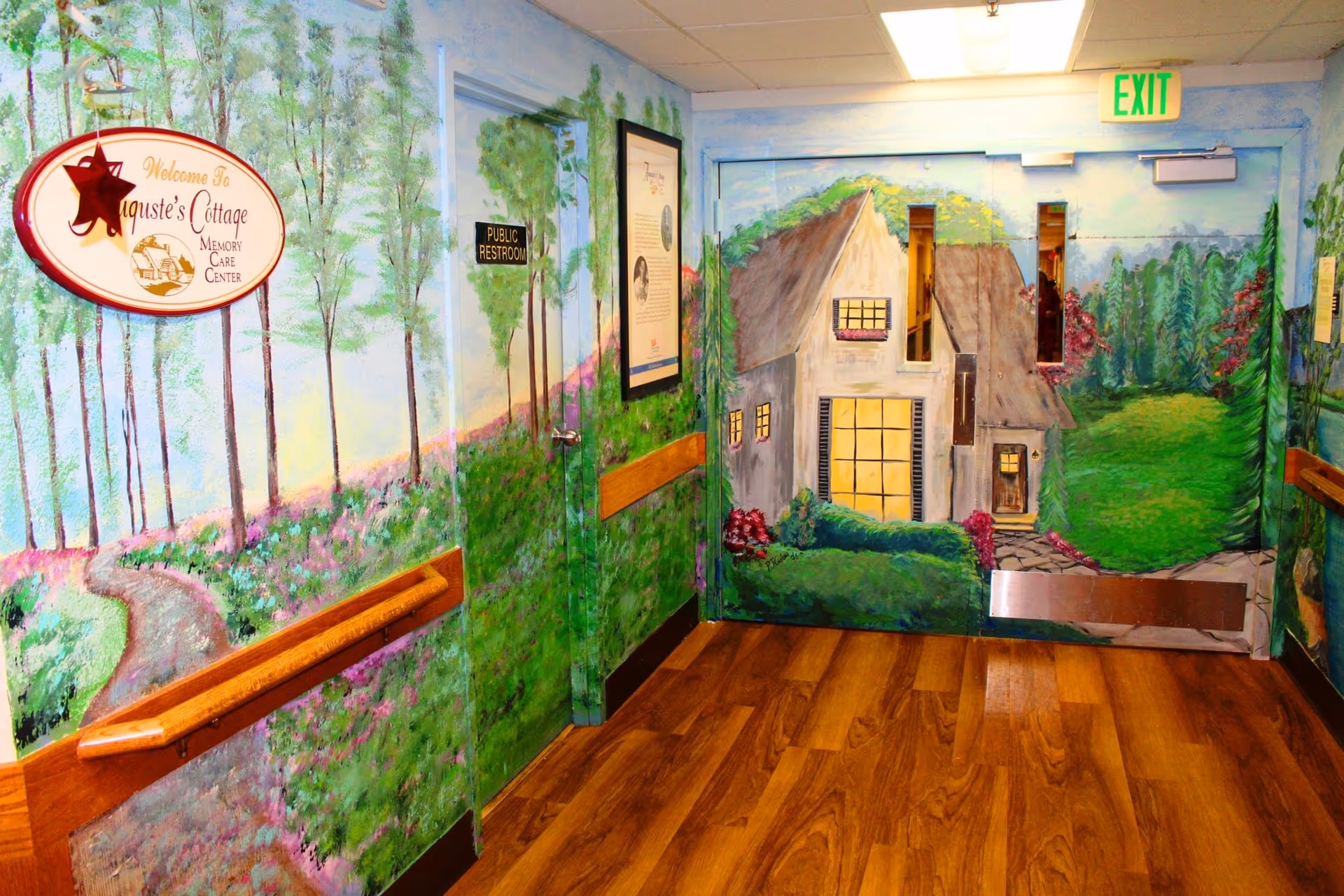 A hallway in a nursing and rehabilitation facility with walls painted in a mural depicting a scenic outdoor landscape including trees, flowers, and a cottage. There are wooden handrails along the walls, a door labeled 'Public Restroom,' and an exit door with windows showing the continuation of the mural. The floor is wood-patterned.