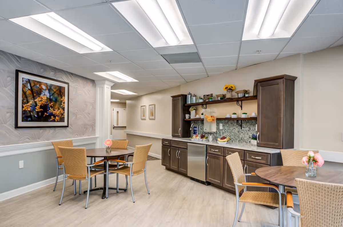 A bright and clean dining area in a senior living facility with two round tables surrounded by wicker chairs. The room features a kitchenette with dark wood cabinets, a small refrigerator, a sink, and a backsplash with mosaic tiles. There are decorative items and a pitcher of infused water on the counter. The walls are light-colored with a framed picture of autumn leaves and soft overhead lighting.