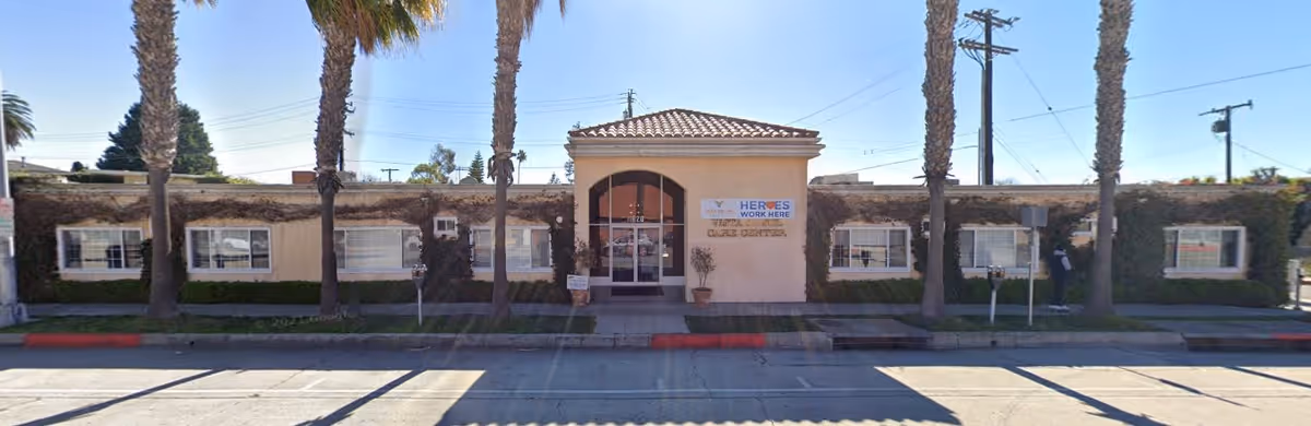 Front exterior view of Vista Del Sol Care Center, a single-story building with a tiled roof, several windows, and palm trees lining the sidewalk in front. There is a sign near the entrance that reads 'Heroes Work Here'.