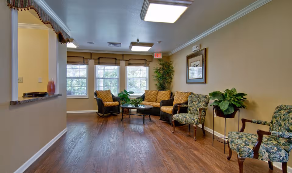 A cozy assisted living facility common area with wooden flooring, several cushioned chairs and a loveseat arranged around a glass coffee table, large windows with blinds letting in natural light, potted plants, and framed artwork on the wall.