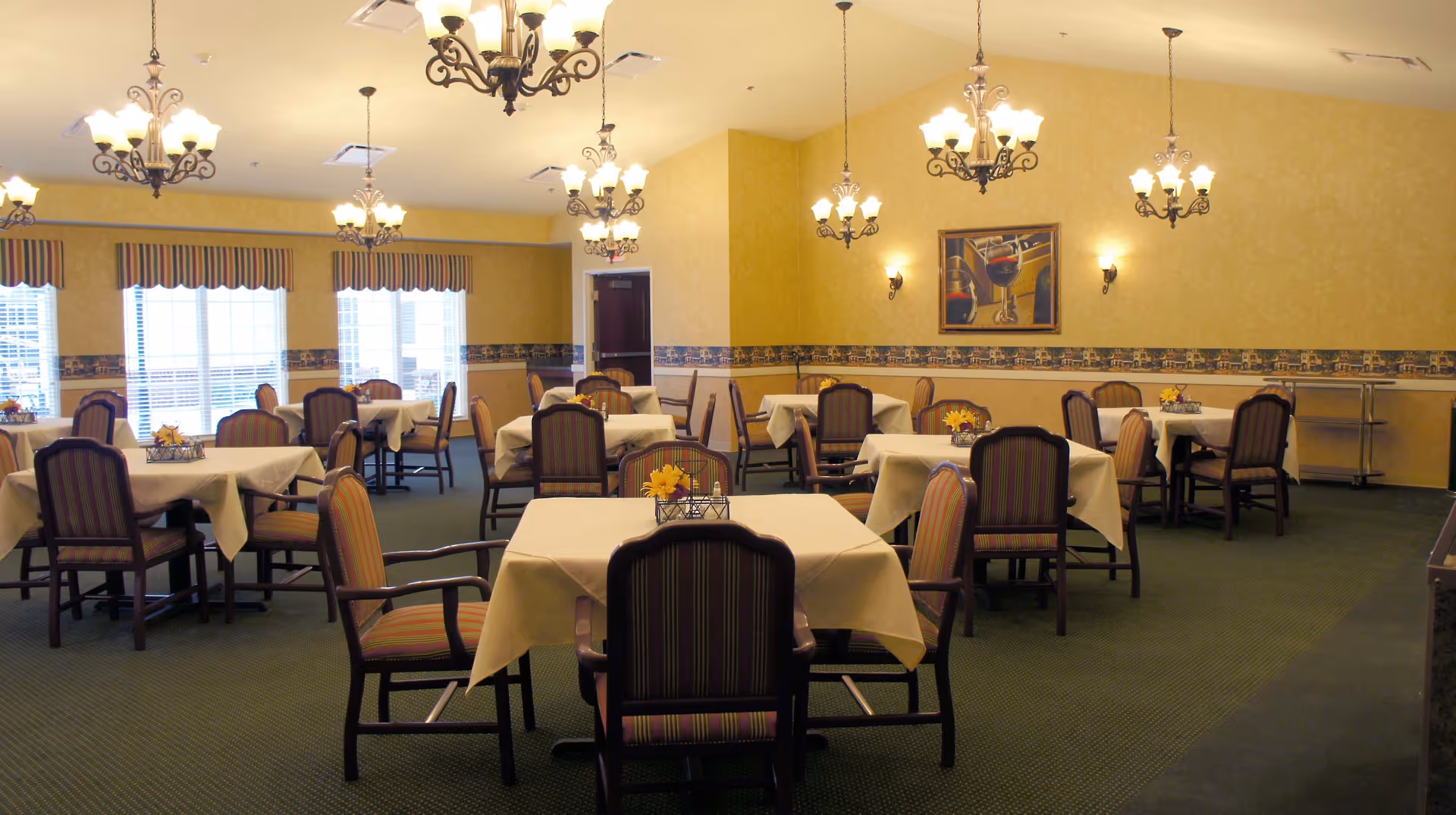 A dining room with multiple tables covered with beige tablecloths and surrounded by wooden chairs with striped upholstery. The room has yellow walls with a decorative border and several chandeliers hanging from the ceiling. There are large windows with striped valances letting in natural light and a painting on the wall depicting wine glasses.