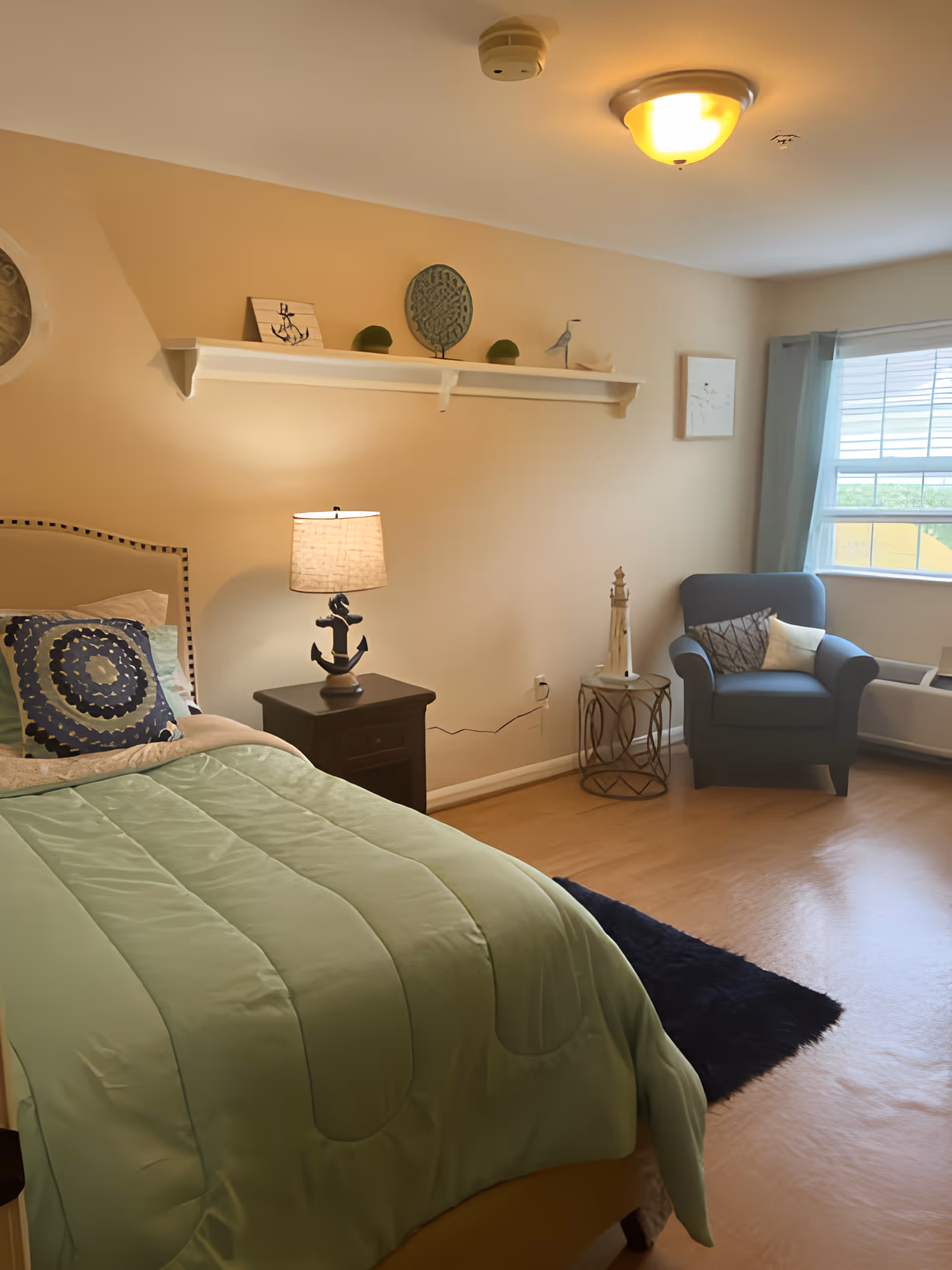 A cozy bedroom with a single bed covered in a green comforter and decorative pillows. Next to the bed is a dark wooden nightstand with a lamp featuring an anchor-shaped base. Above the nightstand is a white wall shelf with nautical-themed decorations including a small anchor artwork, a round green decorative piece, and a bird figurine. In the corner near a window with light blue curtains, there is a blue armchair with two pillows. The room has light-colored walls and flooring, with a small dark rug partially visible on the floor.