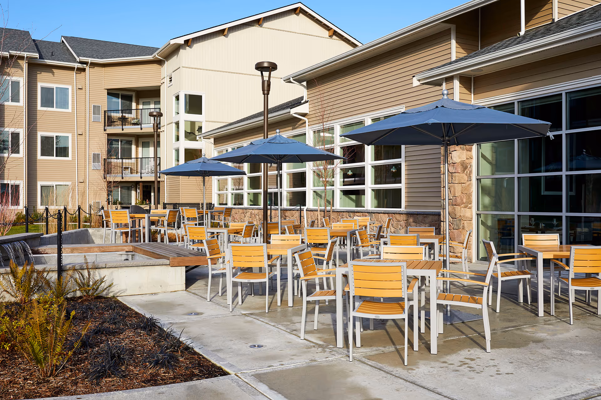 Outdoor seating area with tables, chairs, and umbrellas on a patio beside a multi-story residential building.
