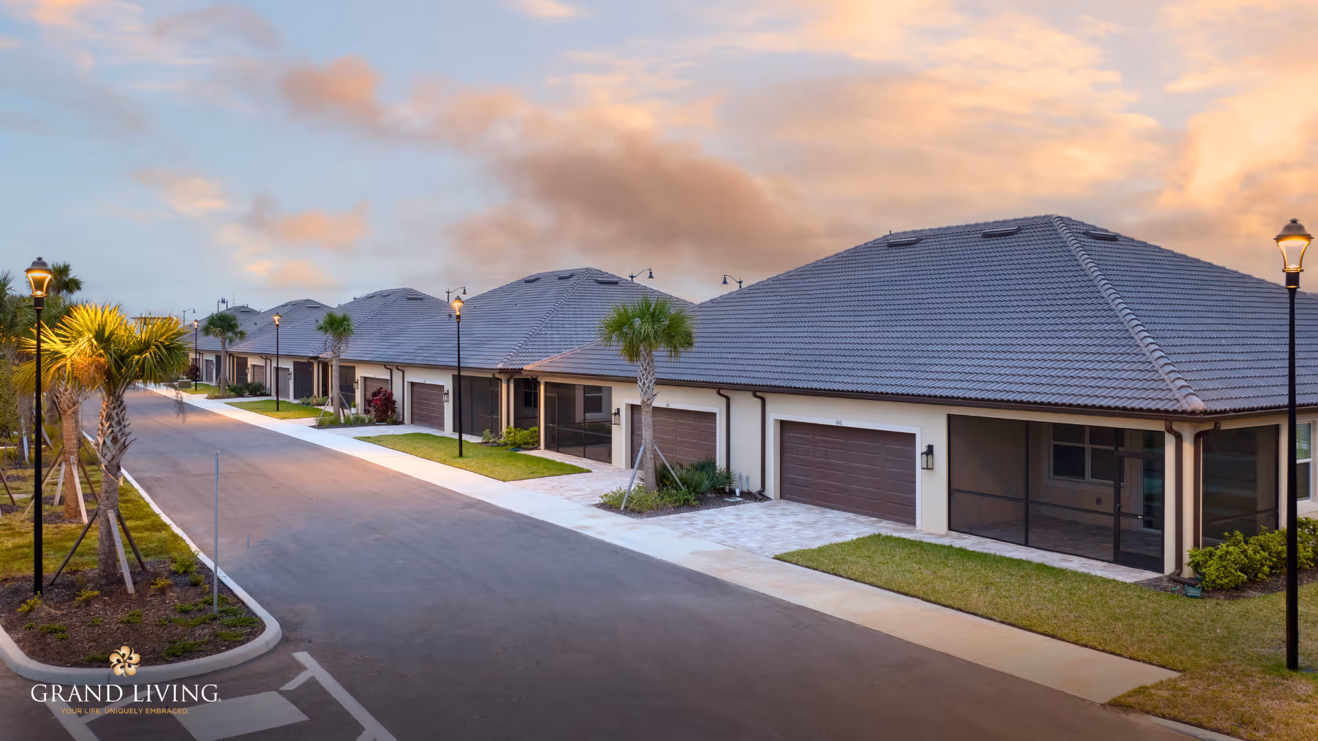 A row of single-story residential buildings with tiled roofs and attached garages along a paved street lined with palm trees and street lamps under a partly cloudy sky at sunset.