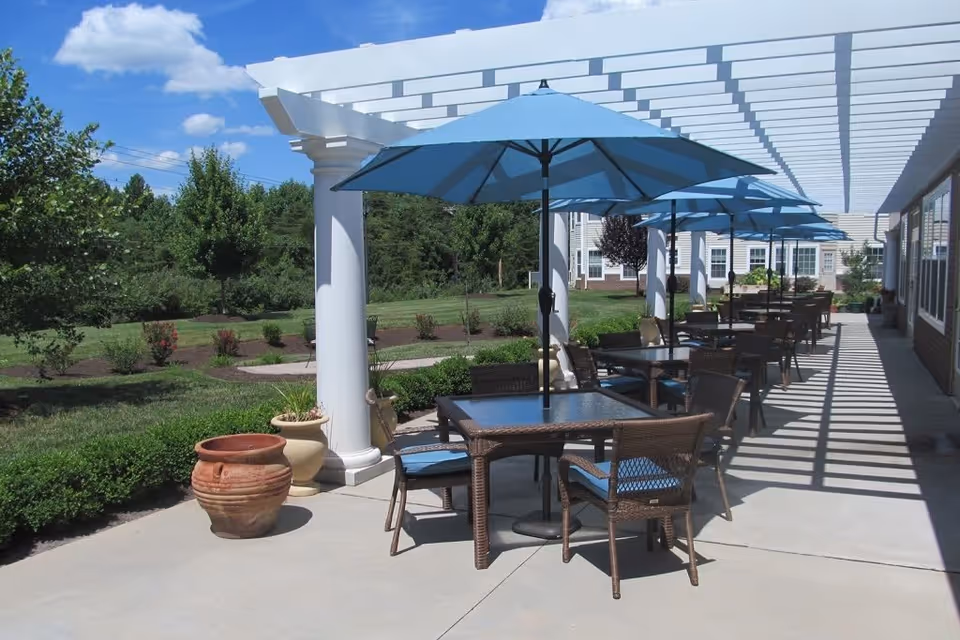 Outdoor patio area with multiple tables and chairs under blue umbrellas, shaded by a white pergola. The patio overlooks a landscaped garden with green grass, bushes, and trees under a blue sky with some clouds.