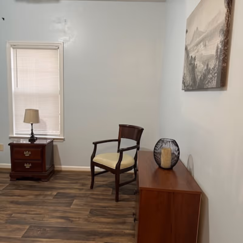 Small furnished room with a wooden chair, nightstand with a lamp, a dresser topped by a decorative vase, a window with blinds, and wall art.