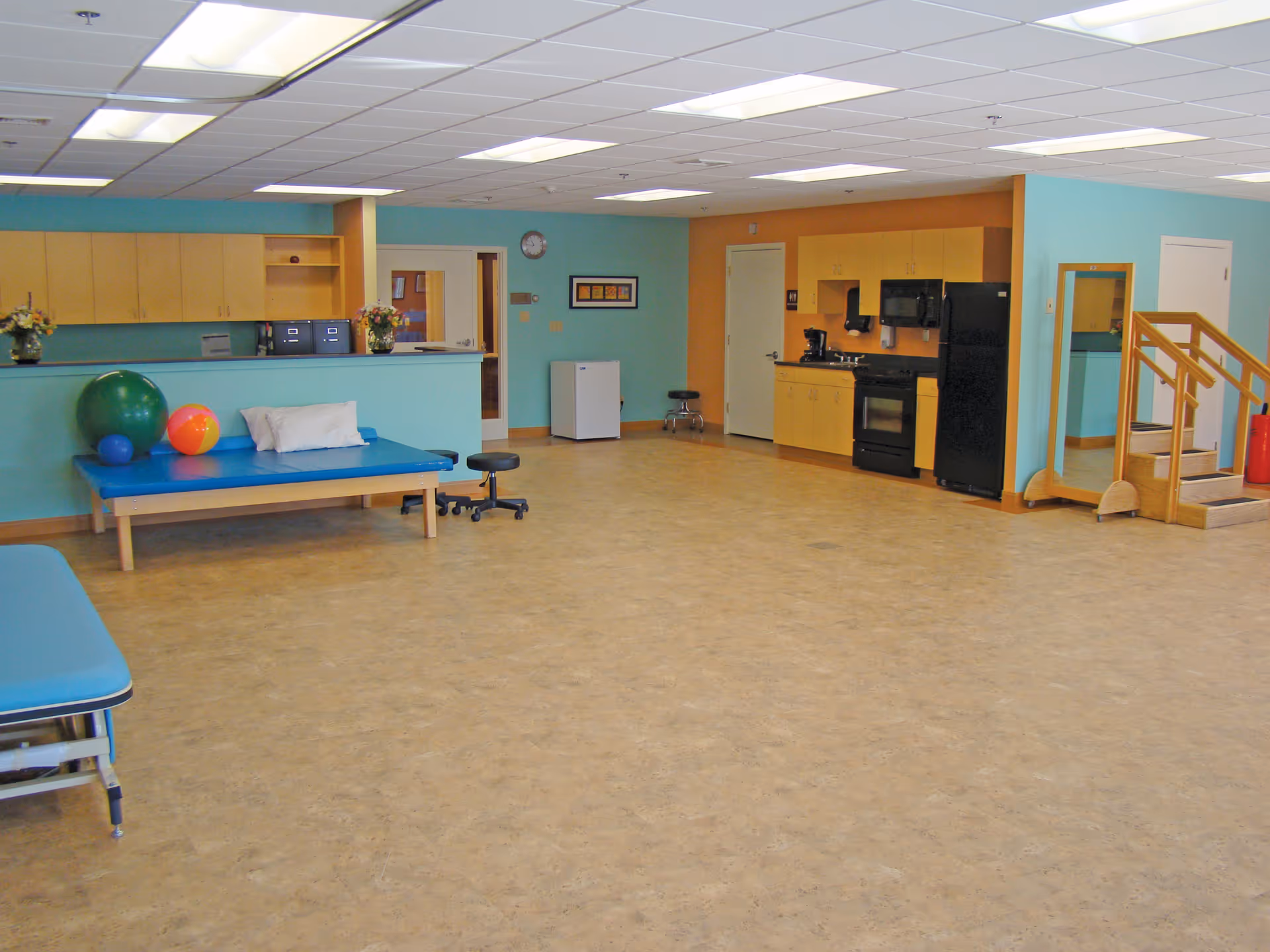 A spacious room with light blue and orange walls, featuring a kitchenette with black appliances, a large mirror, a small set of wooden stairs with handrails, a blue therapy table with exercise balls and a pillow, and a counter with cabinets and flowers.