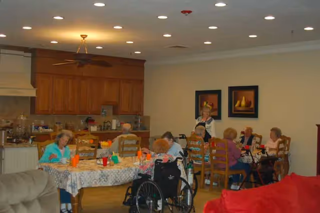 A group of elderly people sitting around two tables in a dining area of an assisted living facility. Some individuals are using wheelchairs and walkers. The room has wooden cabinets, a kitchen area in the background, and framed fruit paintings on the wall.