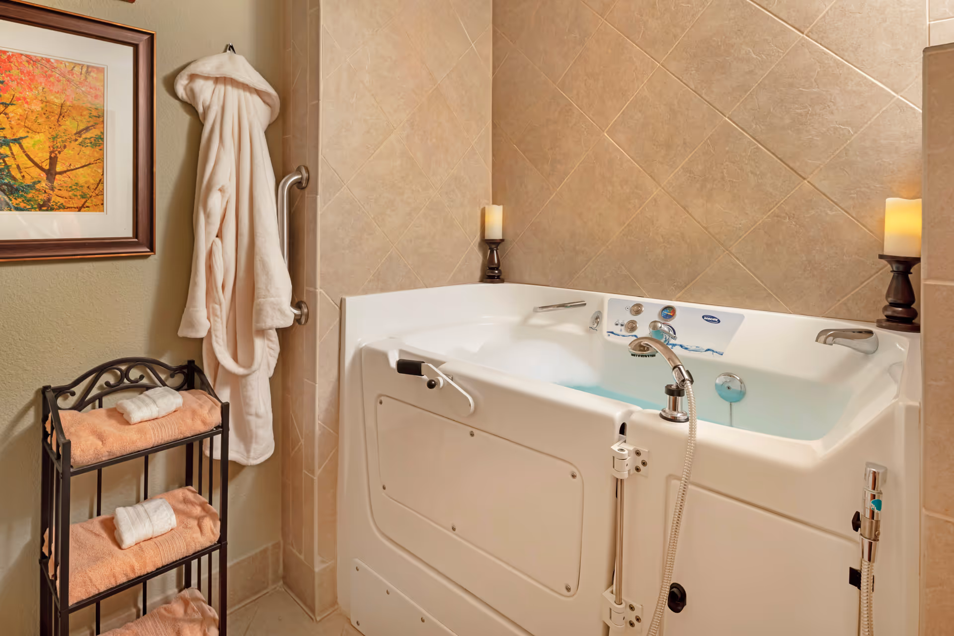 A walk-in bathtub filled with water and bubbles in a tiled bathroom. There are two candles on holders placed on the tub's edge. A white bathrobe hangs on a wall hook next to a small metal rack holding folded peach-colored towels with white rolled towels on top. A framed picture of autumn trees is on the wall.