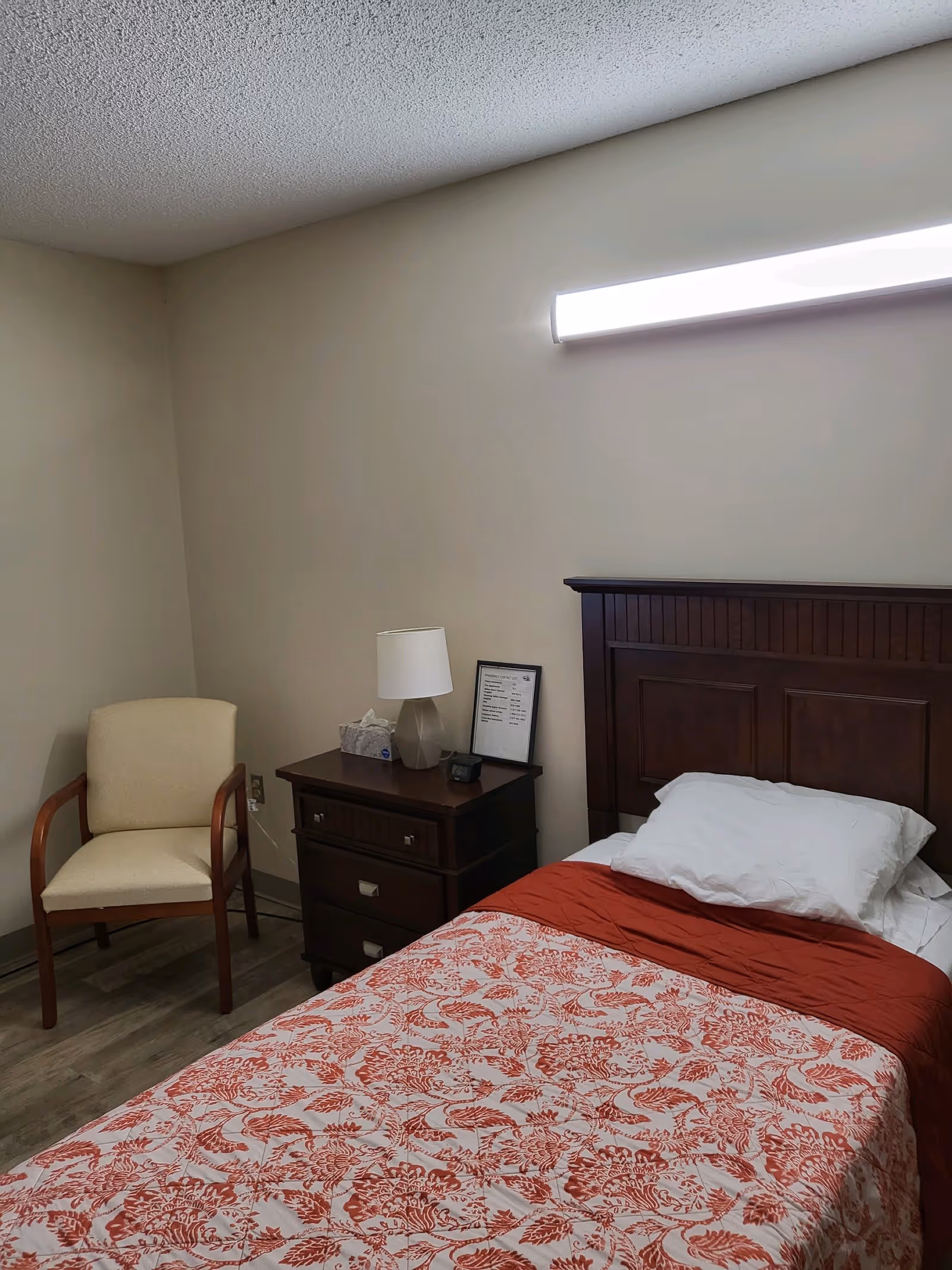 A small bedroom with a single bed covered with a red and white floral patterned bedspread. Next to the bed is a dark wooden nightstand with a white lamp, a box of tissues, and a framed document. There is a beige upholstered chair with wooden armrests placed against the wall. The room has light-colored walls and a wooden floor.