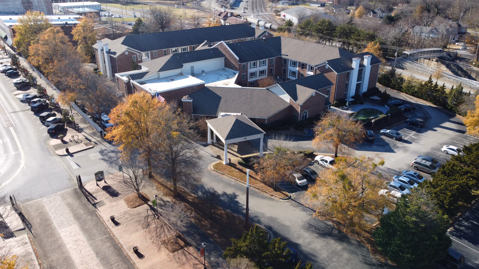 Aerial view of City Center Village Senior Community showing a large multi-wing building with a covered entrance, surrounded by parking lots and trees with autumn foliage. The facility is located near a street with parked cars and sidewalks.