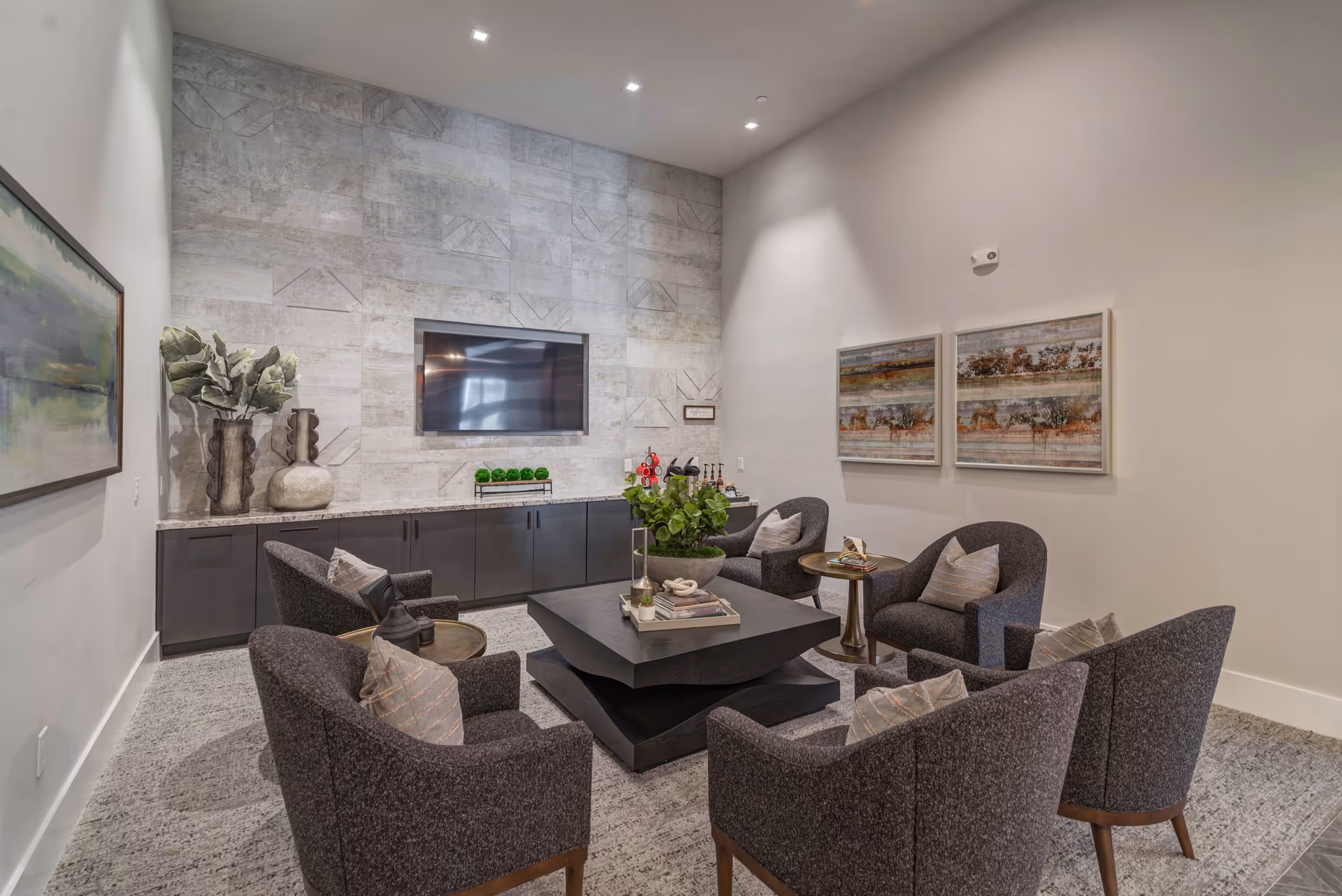 A modern lounge area with six dark upholstered armchairs arranged around a square black coffee table. The room features a textured stone accent wall with a mounted flat-screen TV, a long cabinet below the TV with decorative vases and greenery, and two abstract paintings on the adjacent wall. The space is well-lit with recessed ceiling lights and has a neutral color palette.