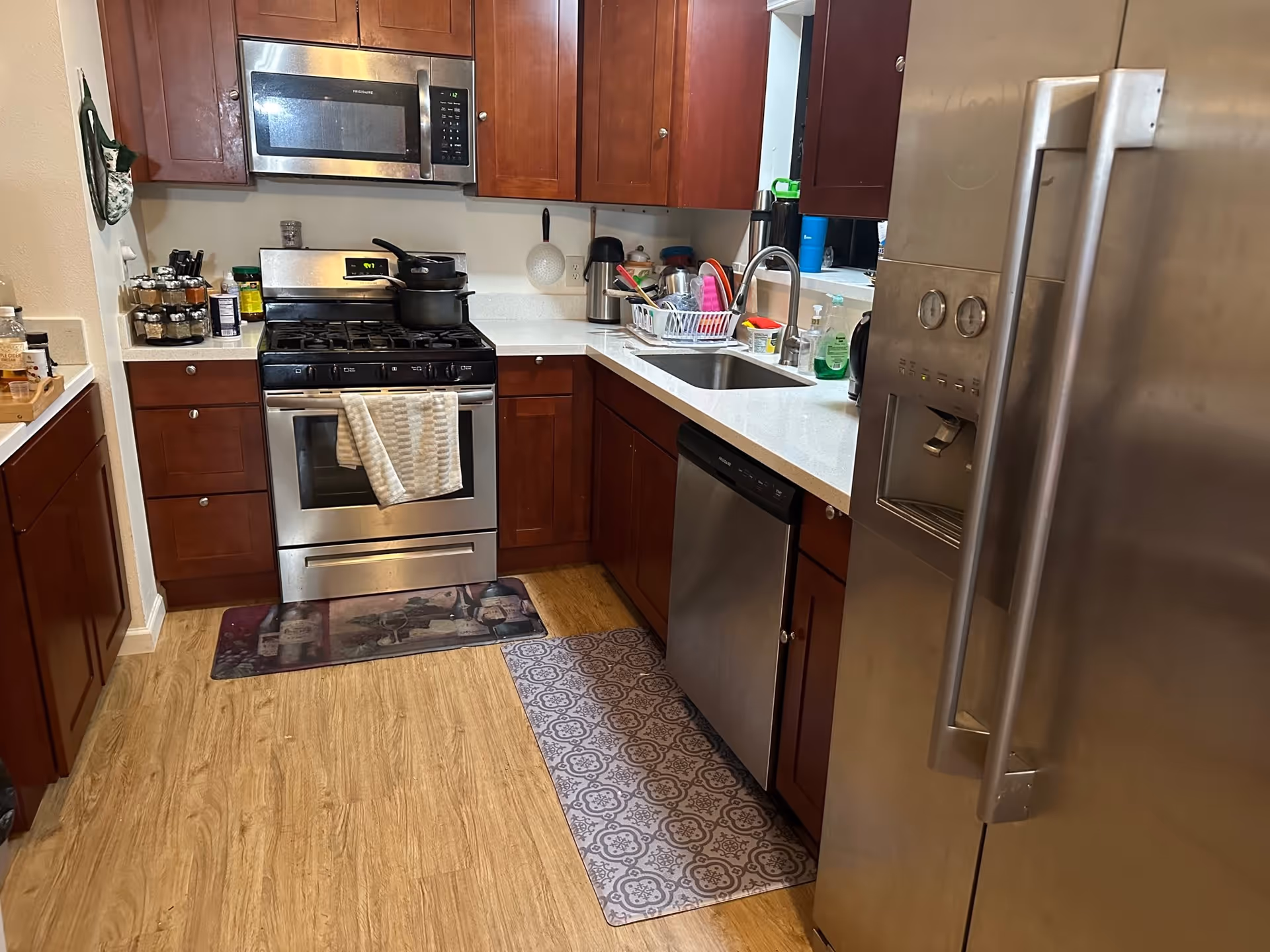 A modern kitchen with wooden cabinets, a stainless steel refrigerator, dishwasher, oven with stove, and microwave. The countertop has a sink with a drying rack filled with dishes and various kitchen items. There are two floor mats in front of the stove and sink area.