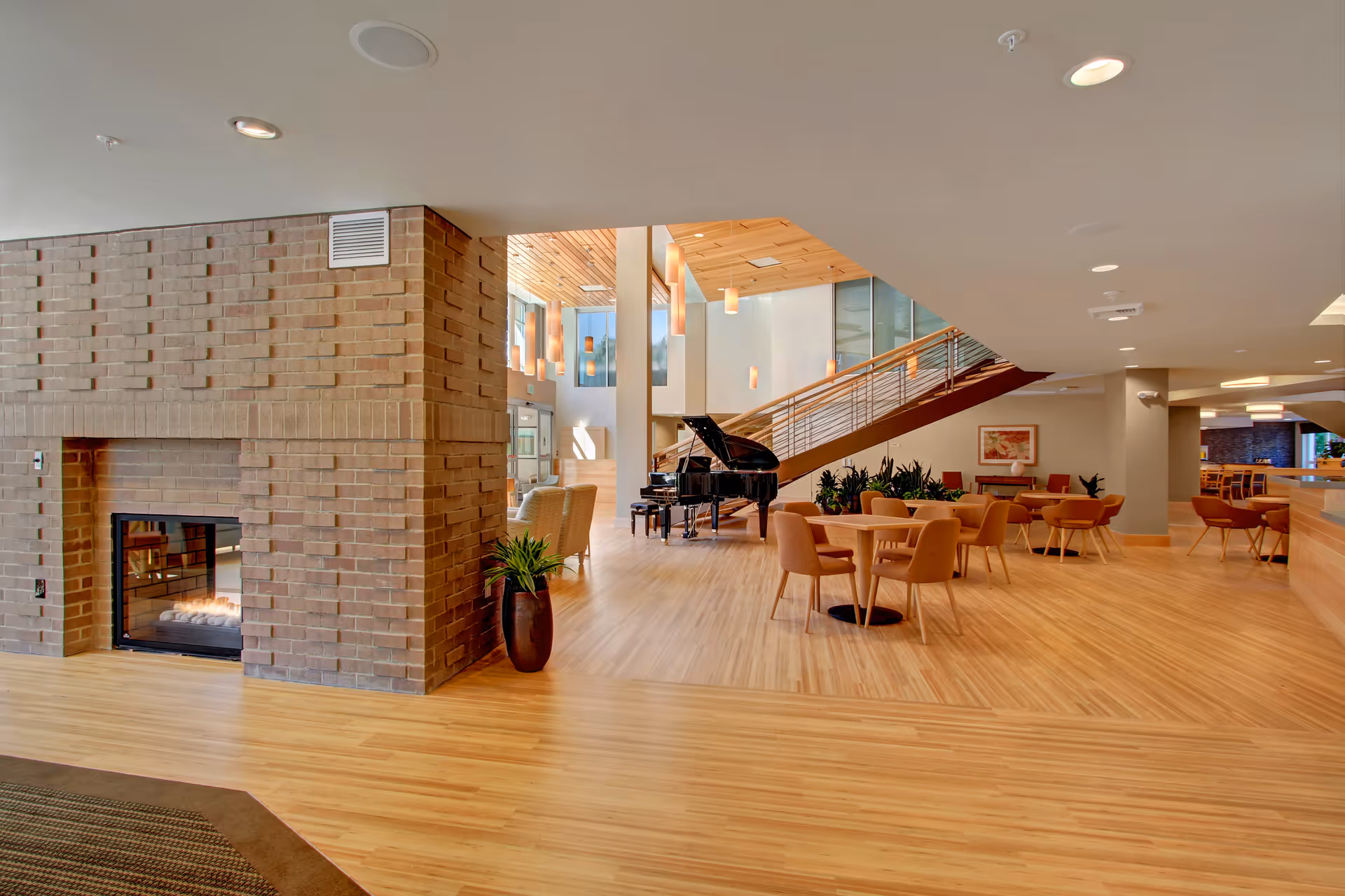 Spacious senior living facility common area with light wood flooring, a brick fireplace, a grand piano, several round tables with orange upholstered chairs, modern hanging pendant lights, and a staircase with wooden steps and metal railing.