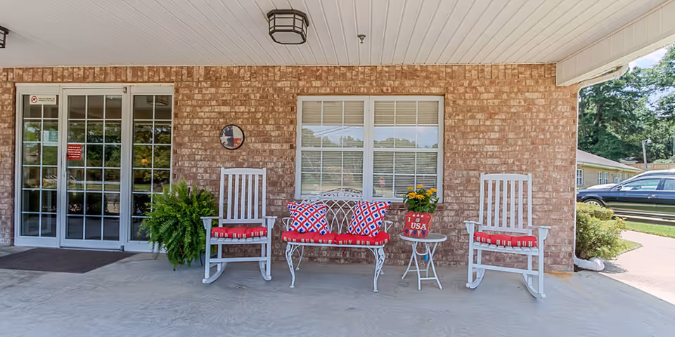 Covered brick front porch showing two white rocking chairs and a bench with red cushions and potted plants outside glass entrance doors.