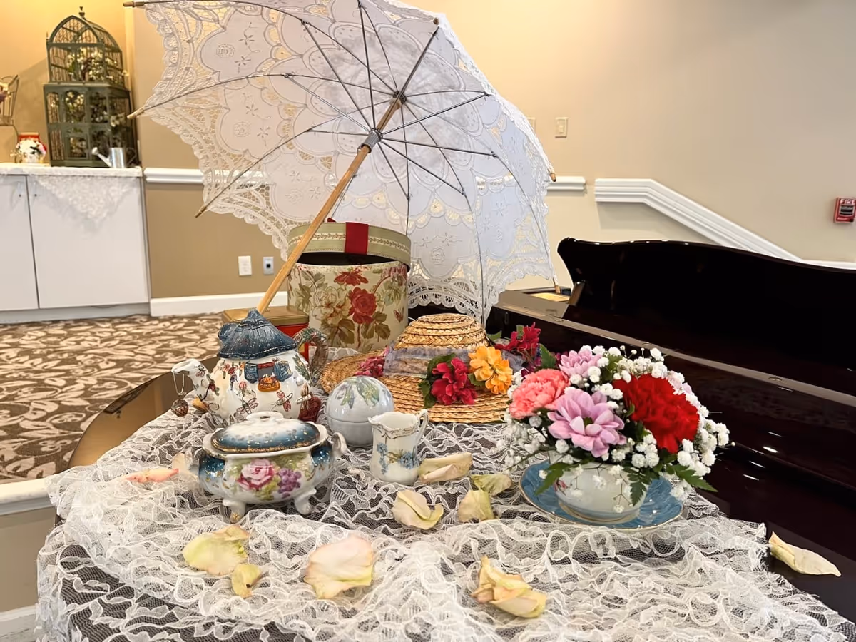 A decorative arrangement on a lace-covered surface featuring a white lace parasol, a floral teapot, a small floral sugar bowl, a creamer, a straw hat adorned with flowers, and a small bouquet of mixed flowers in a teacup. The background shows a carpeted floor, a white cabinet with a lace runner, and a beige wall with a fire alarm.